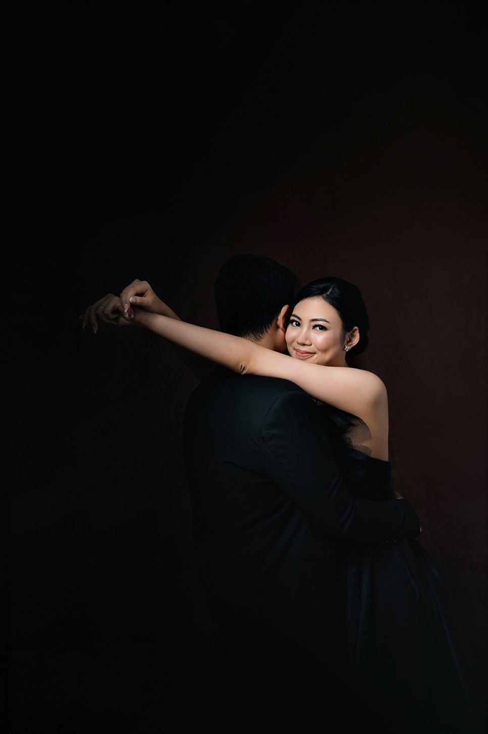 A smiling woman in a black dress dances with a man in black against a dark studio background.