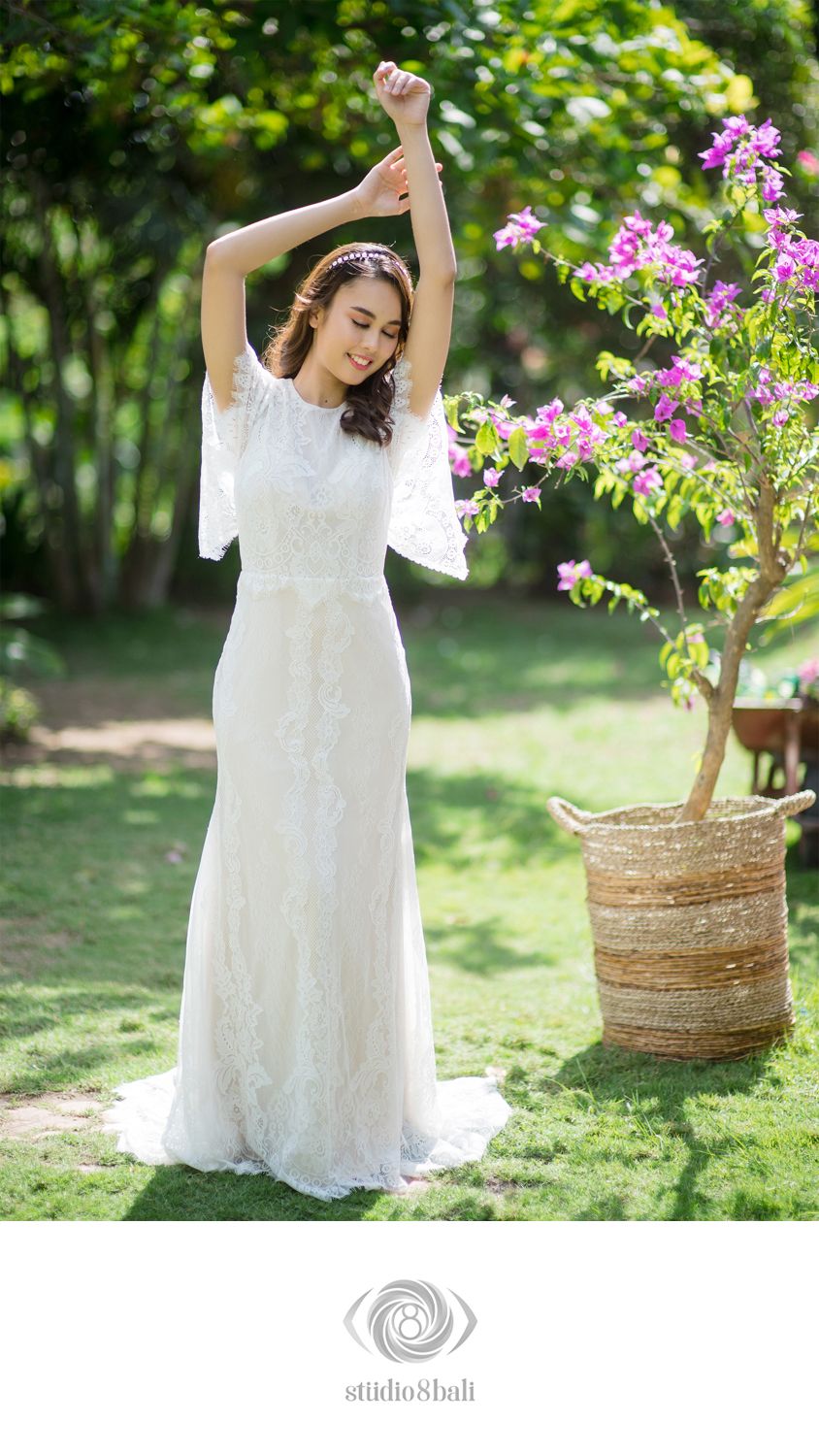 Elegant bride in white lace gown stretching arms overhead beside bougainvillea in sunny garden.