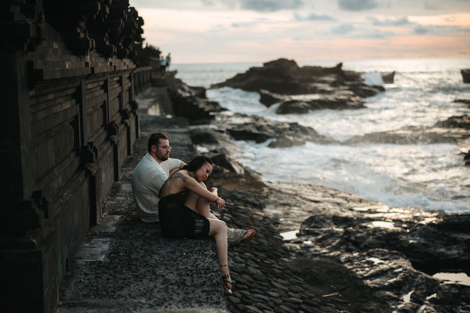 Couple sitting together on rocky coastal steps at sunset, with ocean waves and dramatic cloudy sky in background.