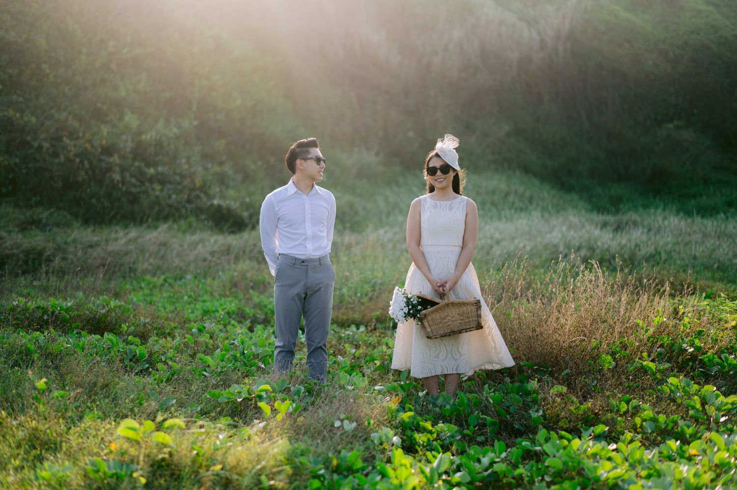 Couple standing in a sunlit green meadow, woman in white dress holding a basket, man in white shirt and gray pants.