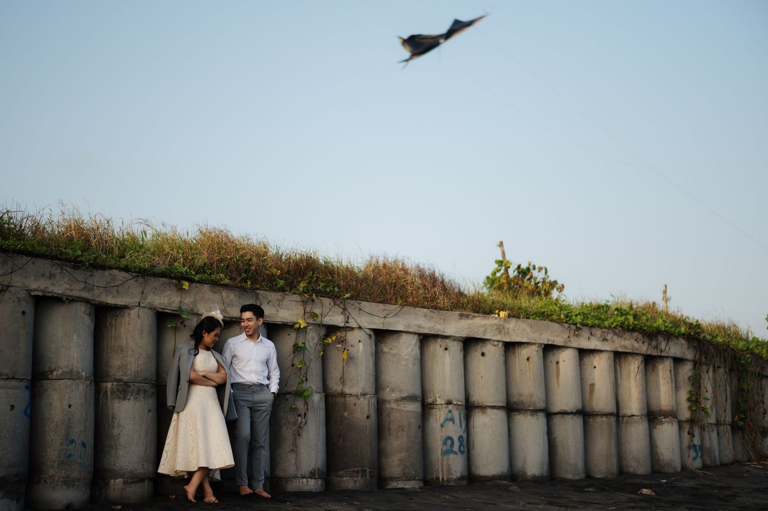 Couple standing by concrete wall with greenery, bird flying overhead against clear blue sky.