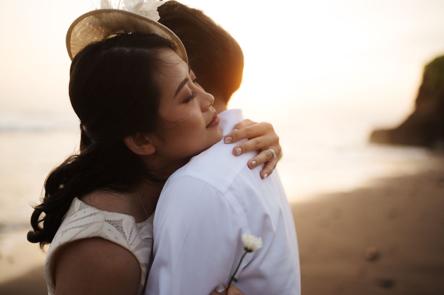 A couple shares a romantic beach hug at sunset, woman in white dress and hat embracing man in white shirt.