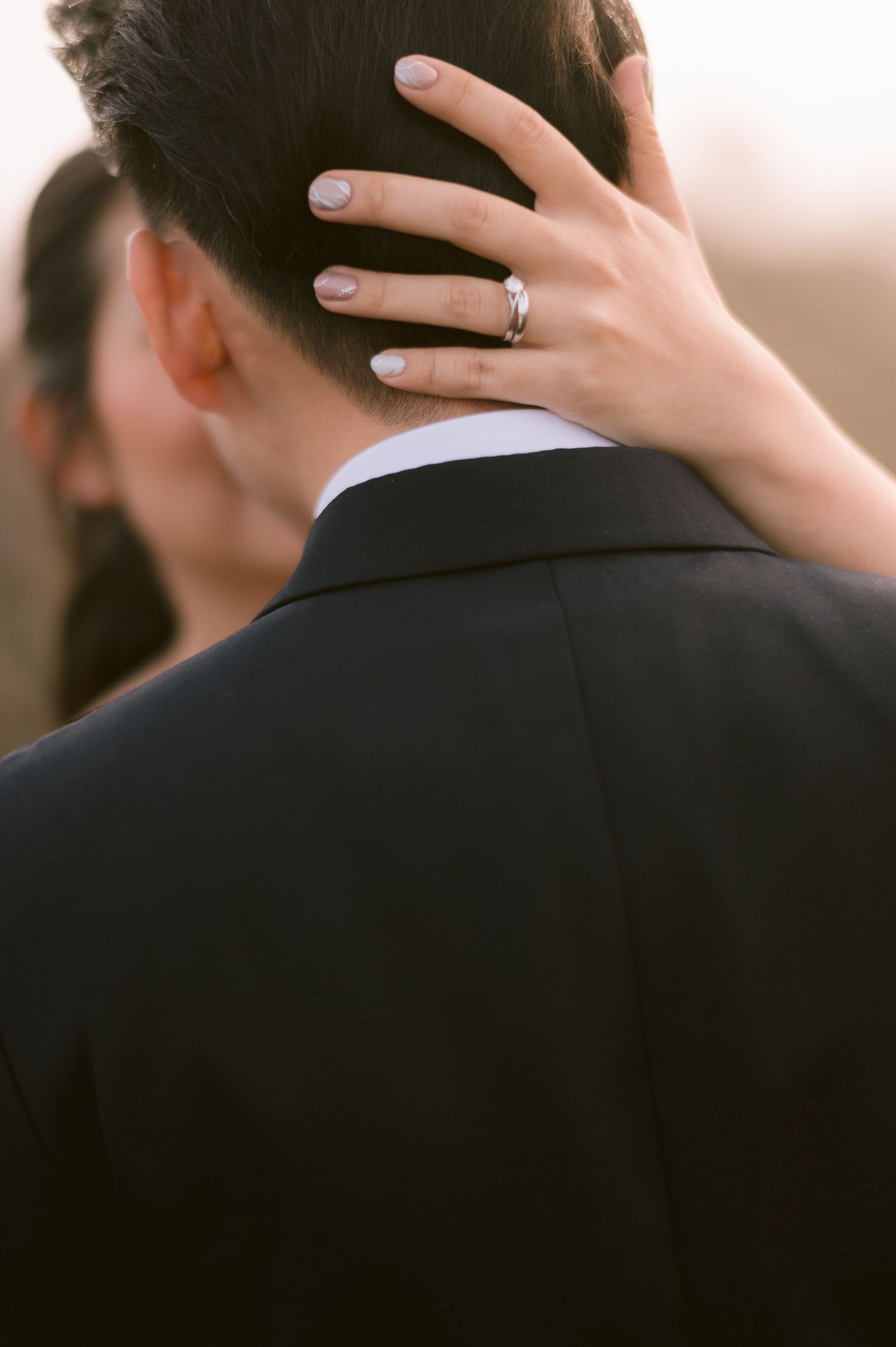 Bride places hand on groom's neck showing engagement ring, couple embracing in elegant black suit at wedding.