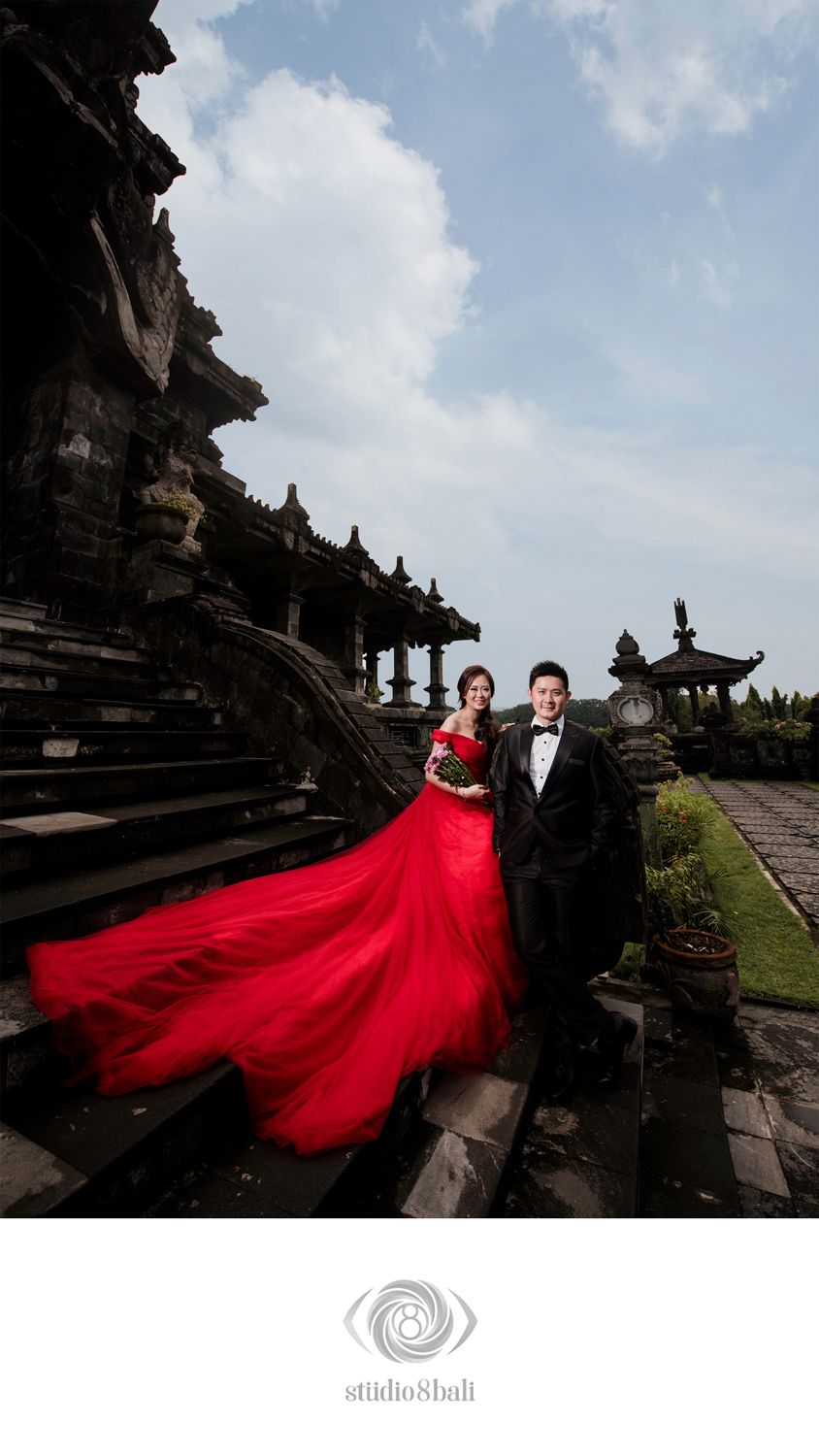 Couple poses at Balinese temple, woman in flowing red gown, man in black tuxedo, dramatic cloudy sky backdrop.