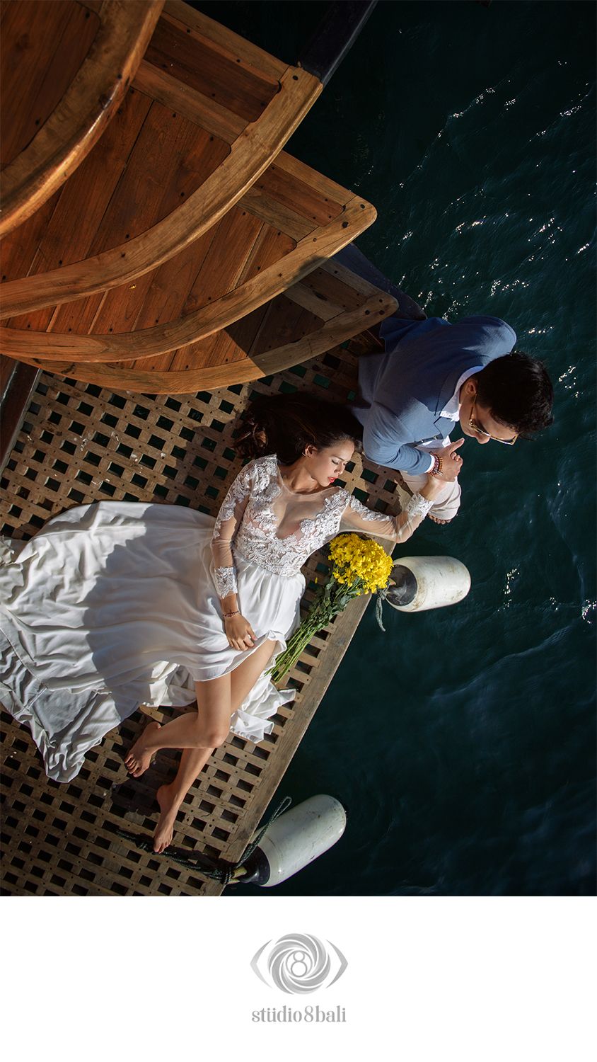 Aerial view of a bride in white lace dress and groom in blue suit on a wooden dock beside a boat over turquoise water.
