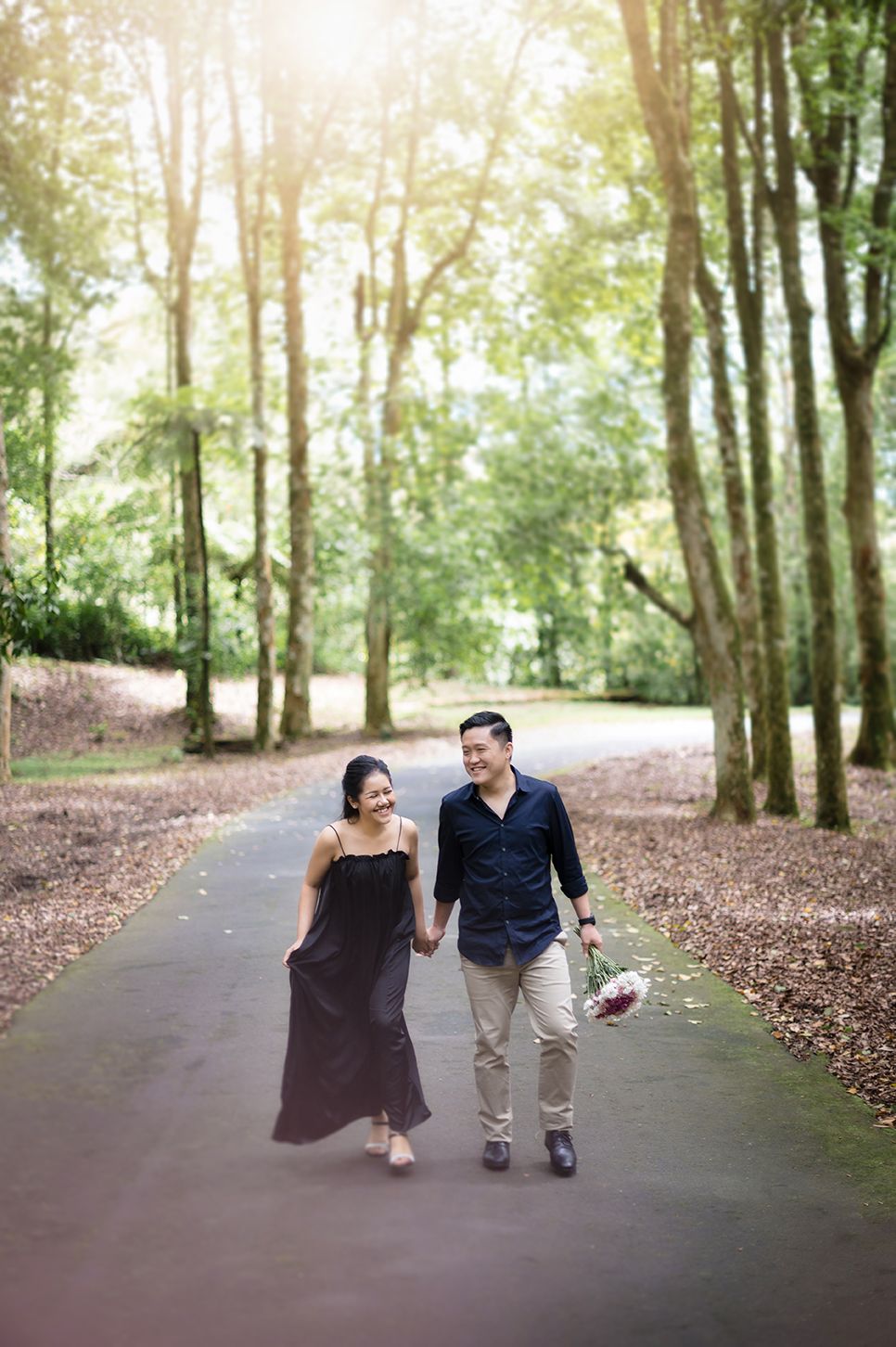 Couple walking hand-in-hand on a sunlit forest path, man holding flowers, woman in black dress, surrounded by tall trees.