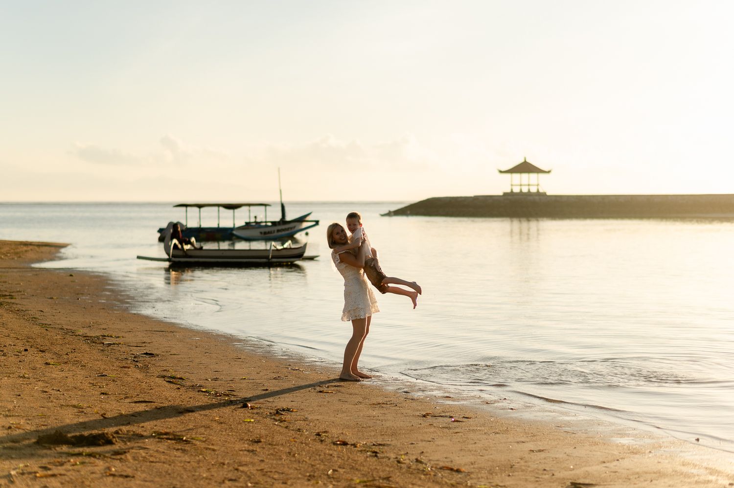 Parent lifting and spinning a child joyfully on a tropical sandy beach during golden hour sunset.