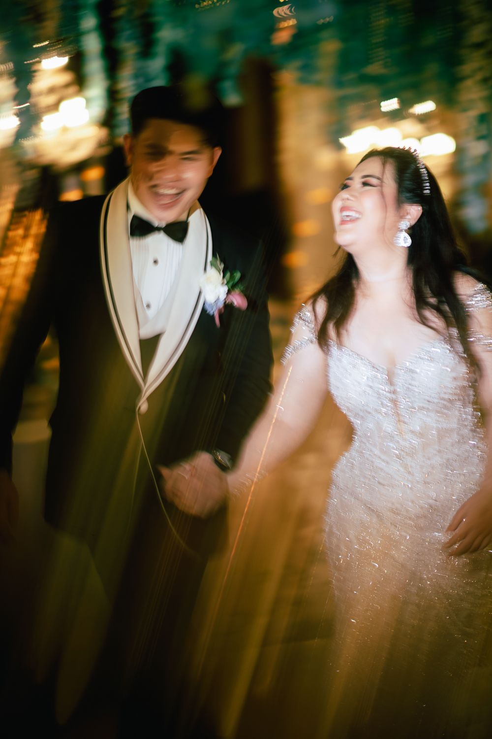 A joyful bride and groom in elegant wedding attire laugh together in a warmly lit venue with a dreamy bokeh effect.