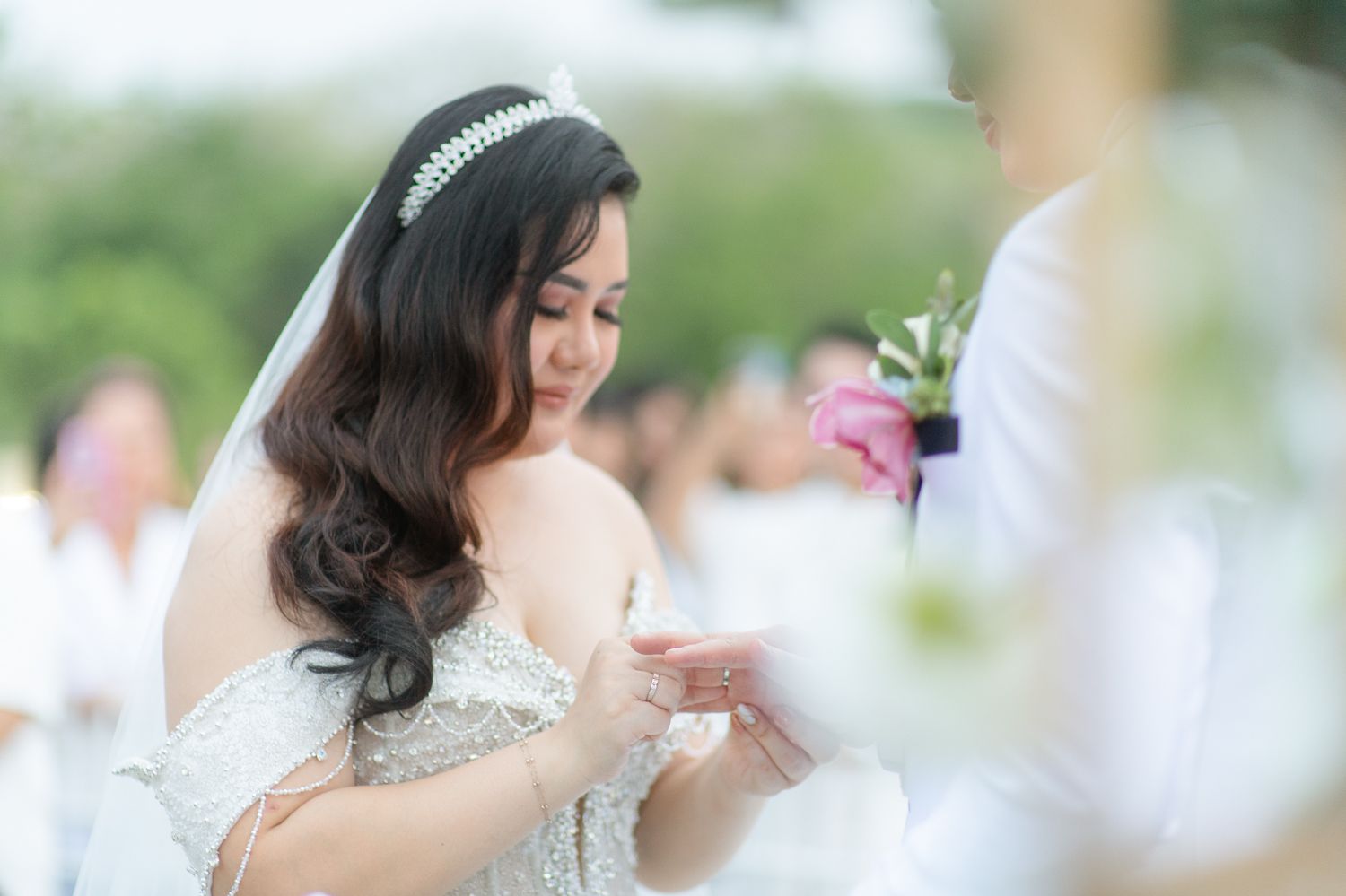 Bride in white gown and tiara placing wedding ring during outdoor ceremony.