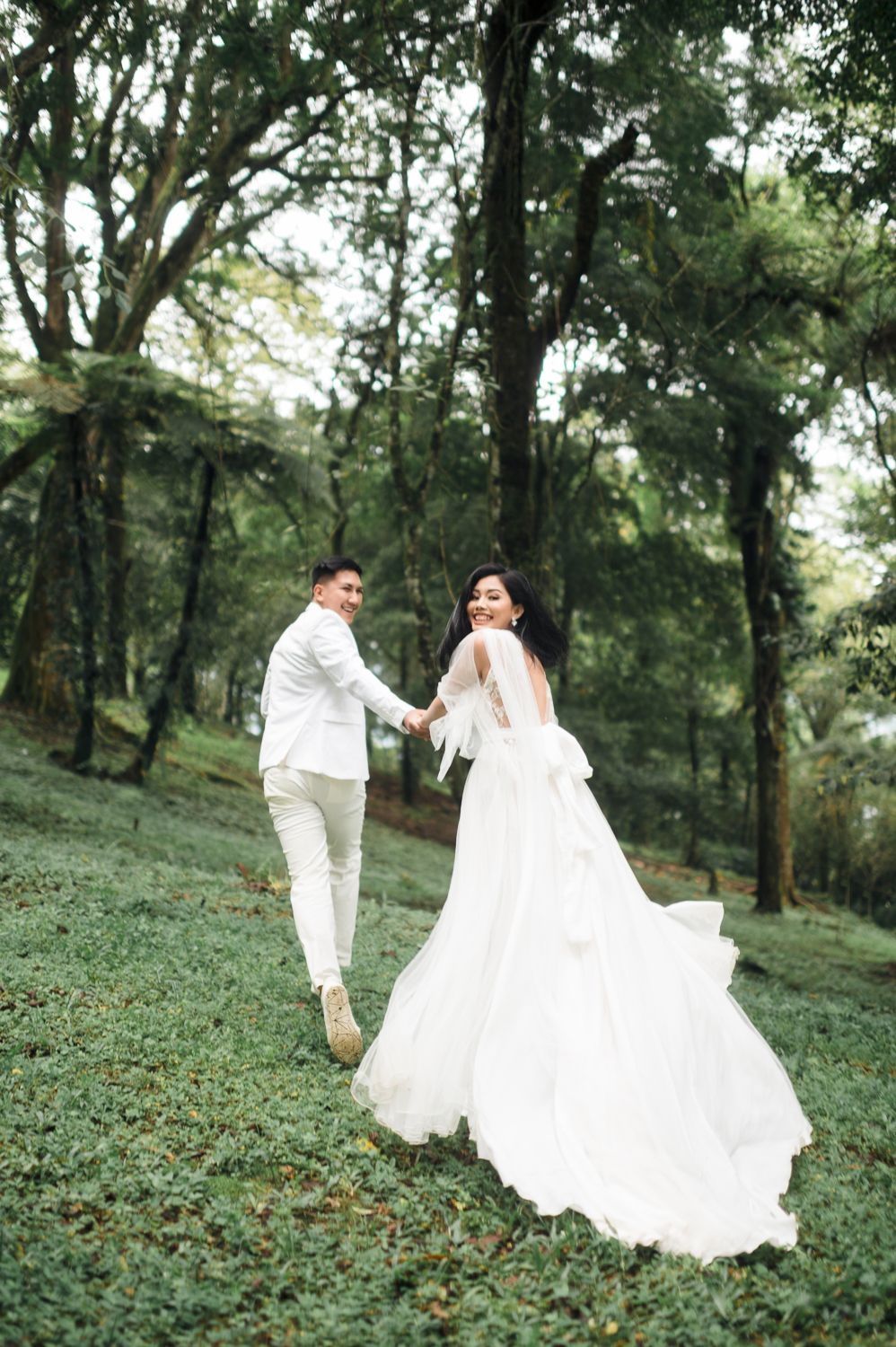 Couple in white wedding attire holding hands and walking through a lush green forest during their outdoor wedding photoshoot.