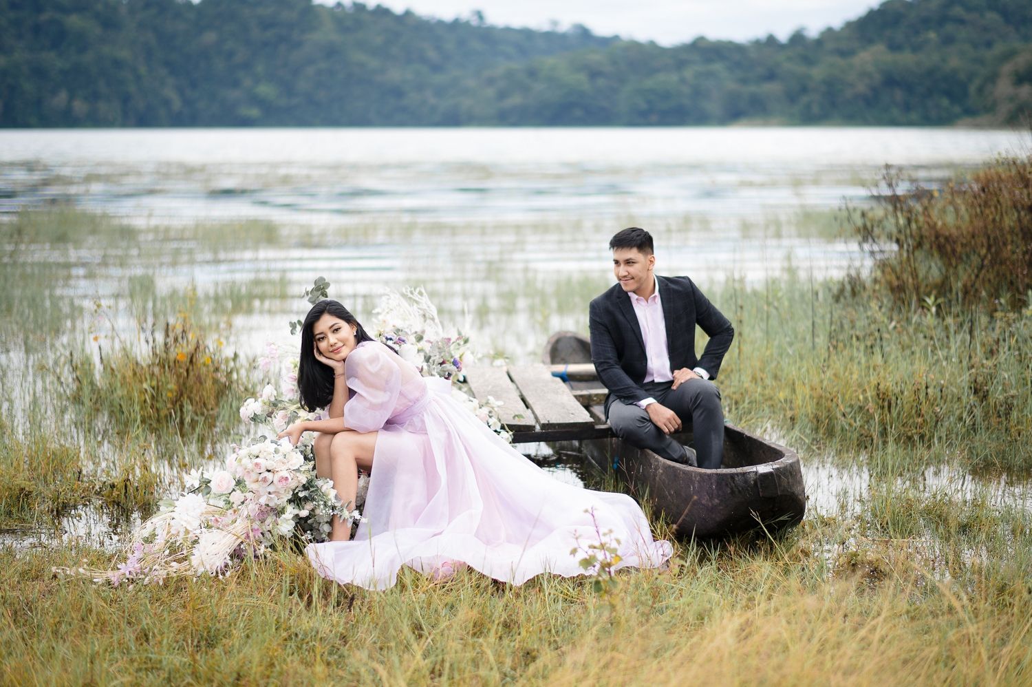 Couple in elegant attire posing in a rustic boat by a serene lake surrounded by lush greenery and floral decorations.