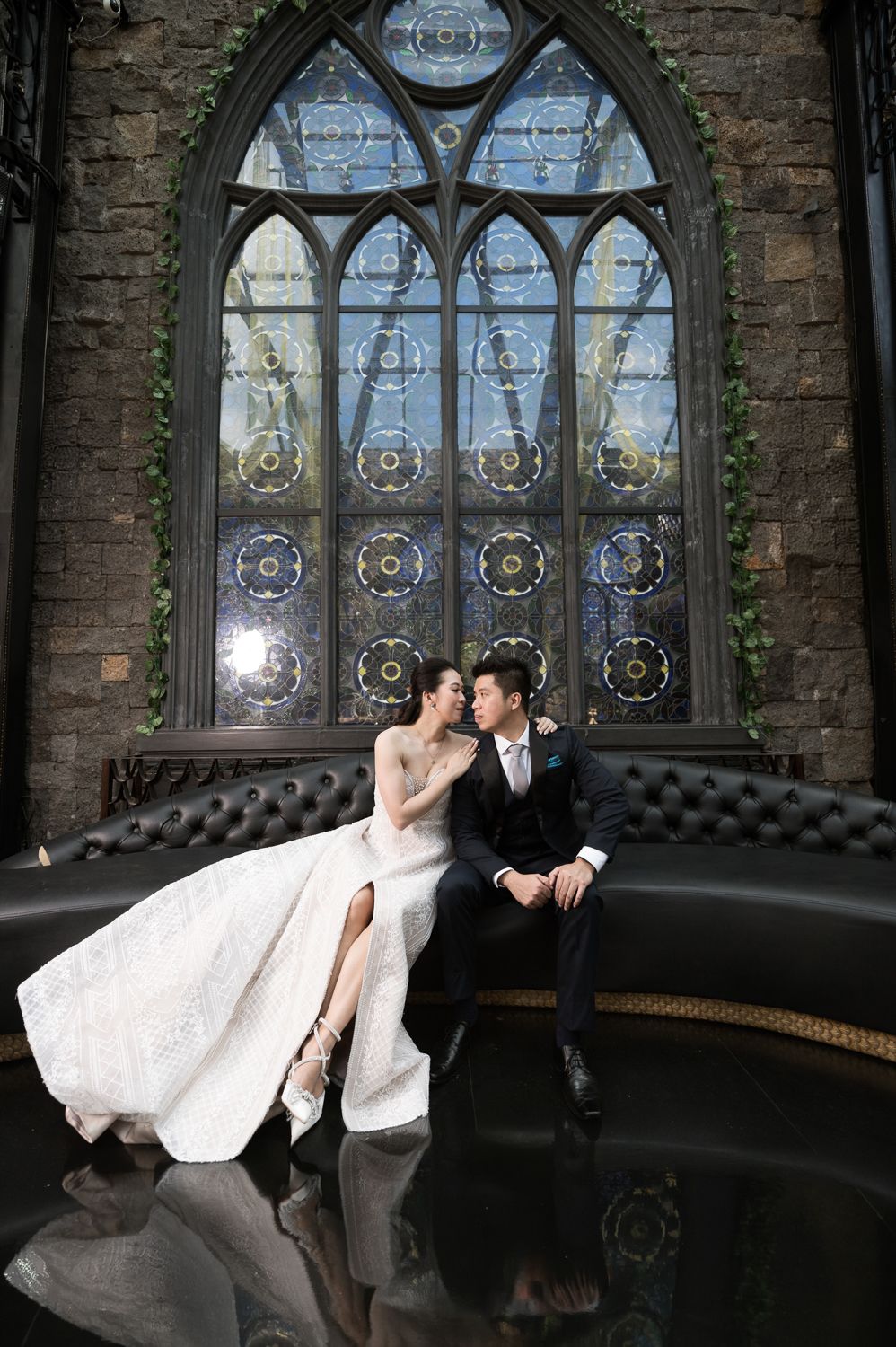 Bride and groom sit together on a tufted sofa before a Gothic stained glass window in an elegant venue.
