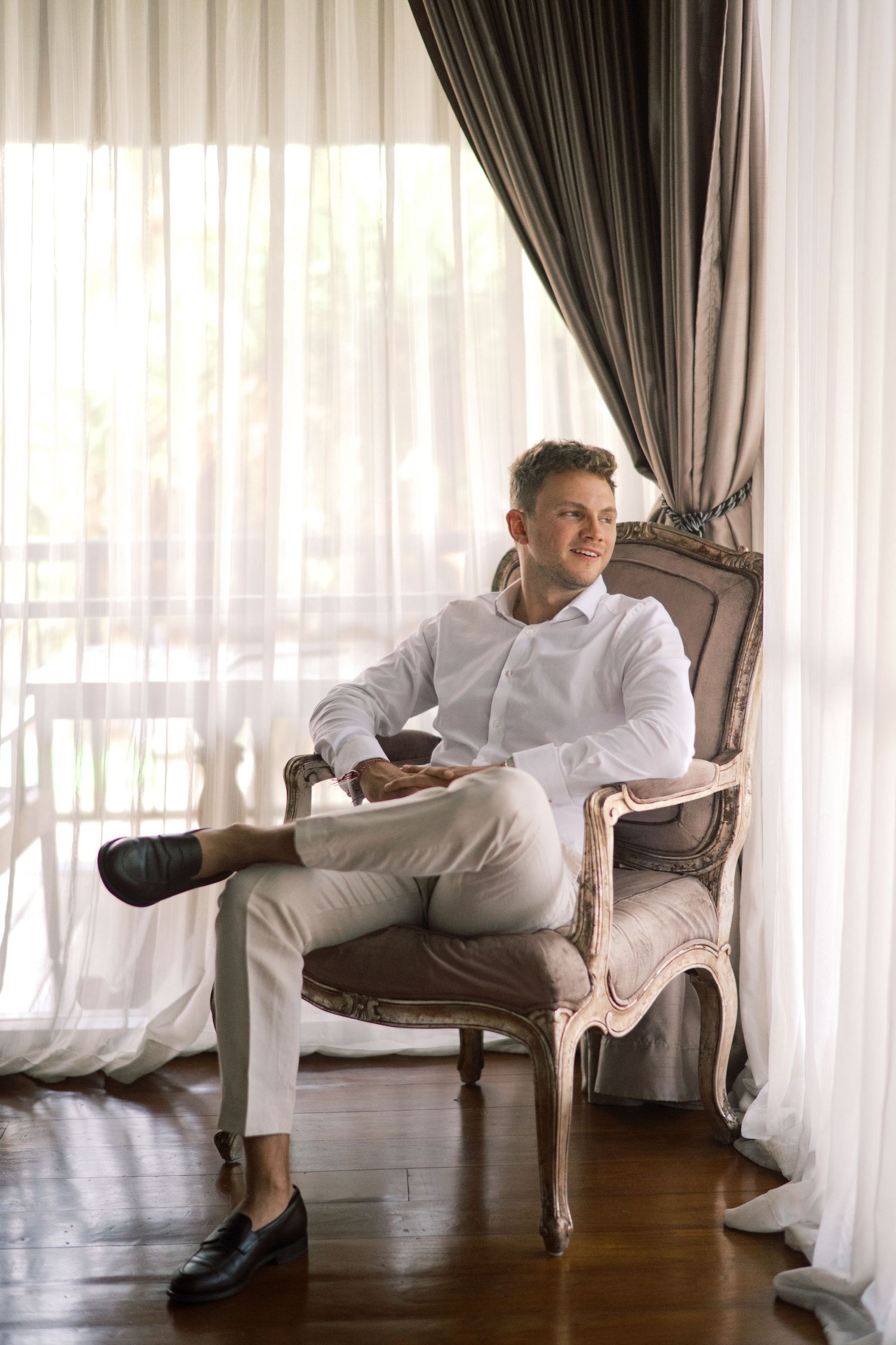 Elegant man in white shirt and beige trousers sitting relaxed in ornate vintage armchair by sheer curtained window.