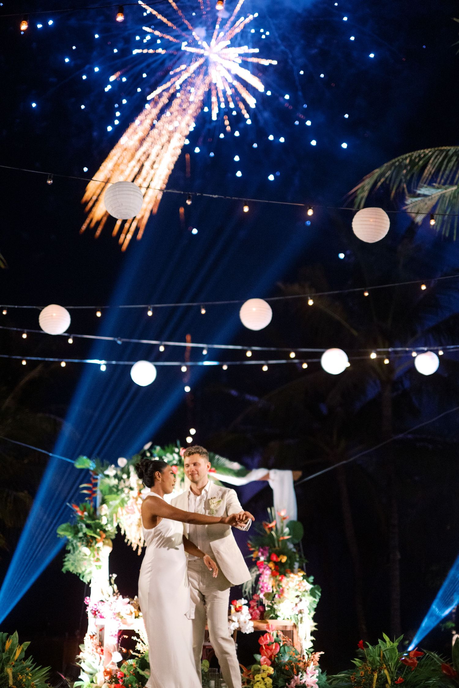 Couple shares first dance under fireworks and string lights at tropical outdoor wedding reception at night.