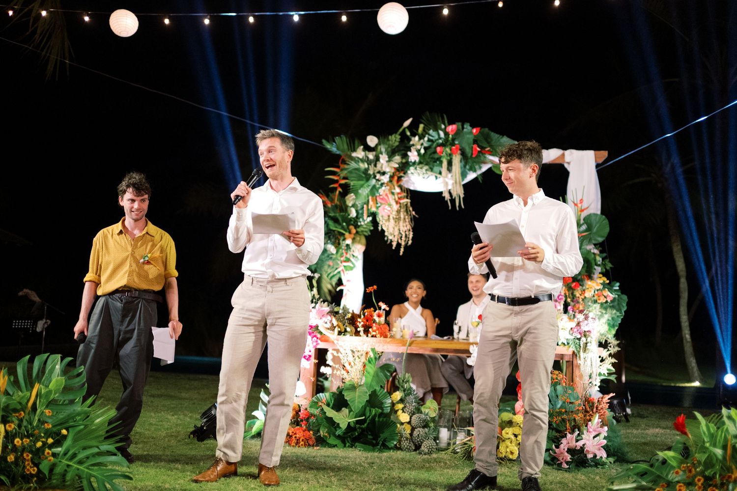 Three men in smart casual attire perform at an outdoor tropical wedding ceremony with floral arch at night.