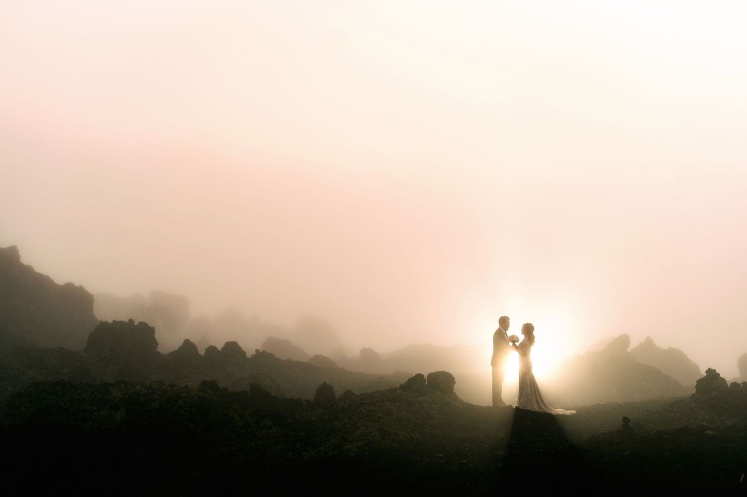 Silhouette of bride and groom embracing in misty golden sunset light on rocky landscape.