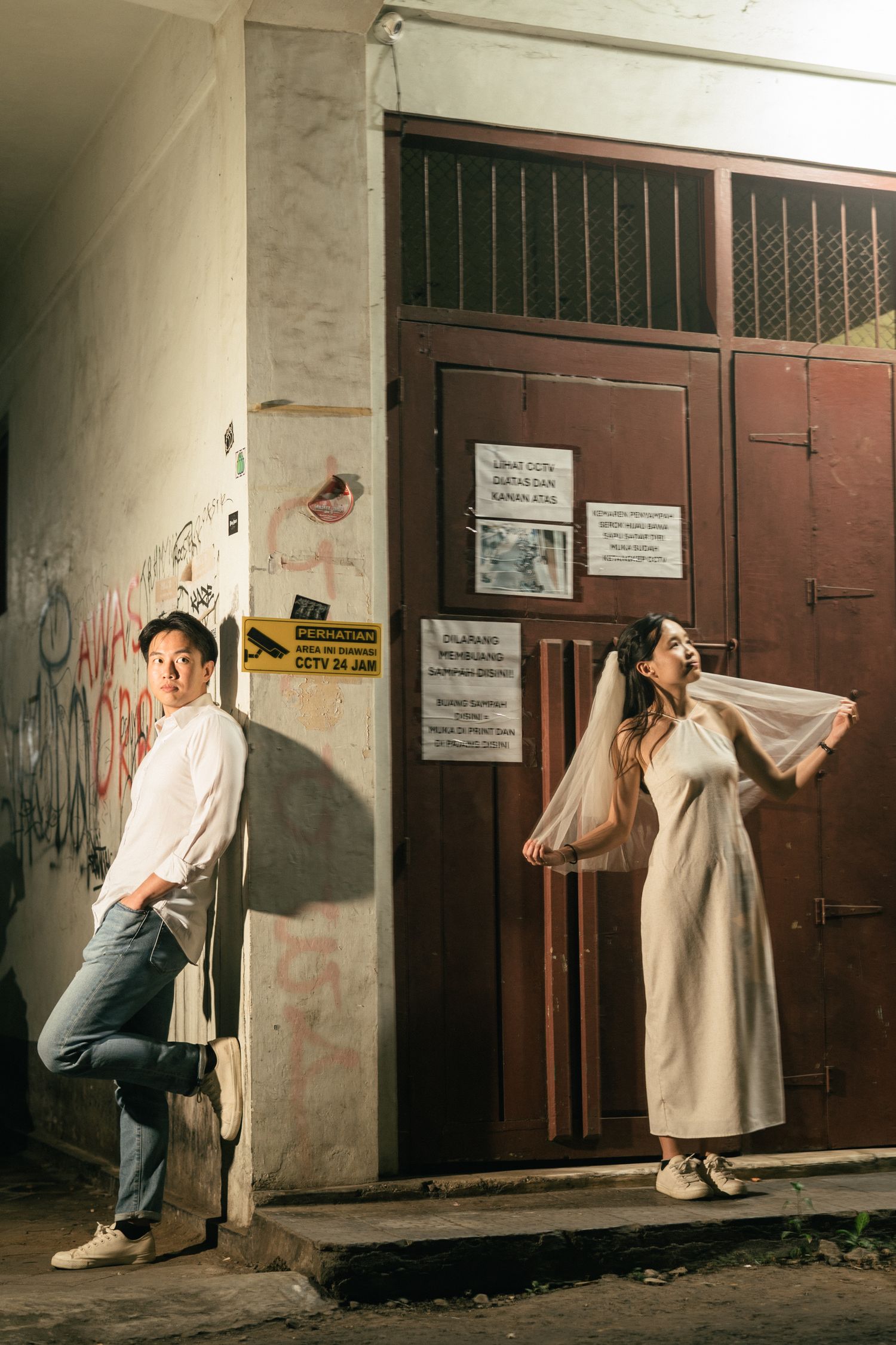 Couple posing outside vintage building with graffiti, man in white shirt and jeans, woman in white dress and veil.