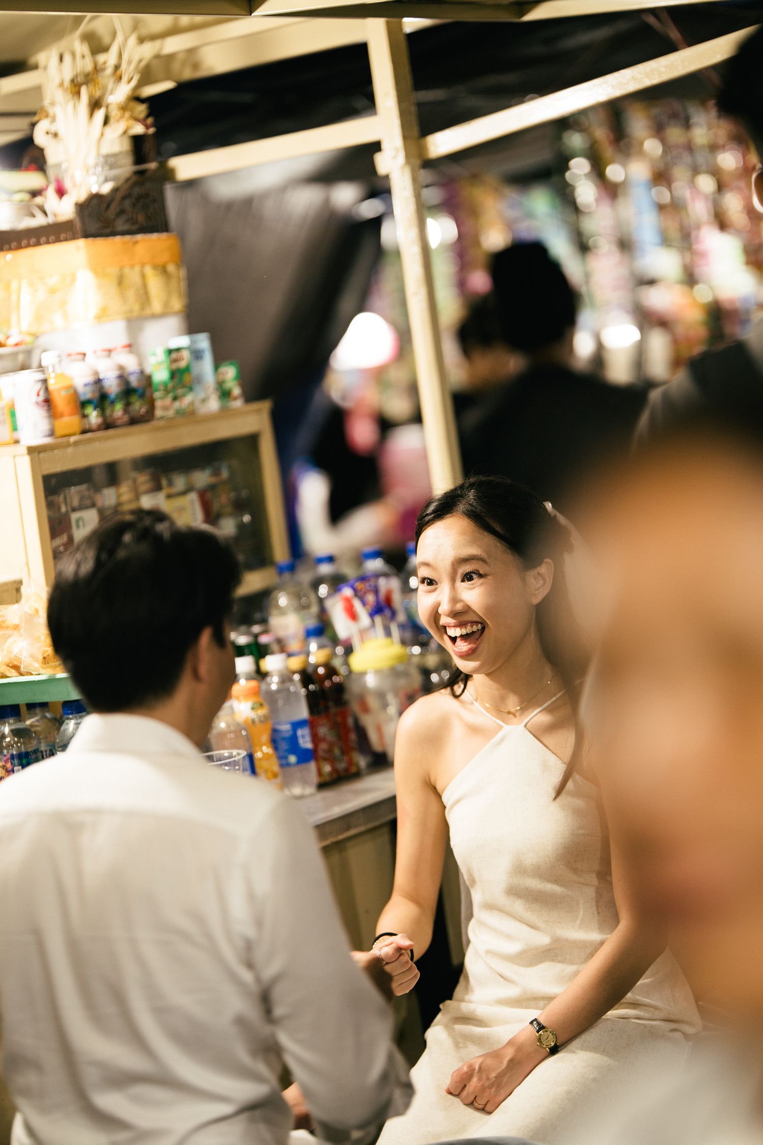 Bride laughing joyfully while holding hands with groom at a vibrant night market stall.