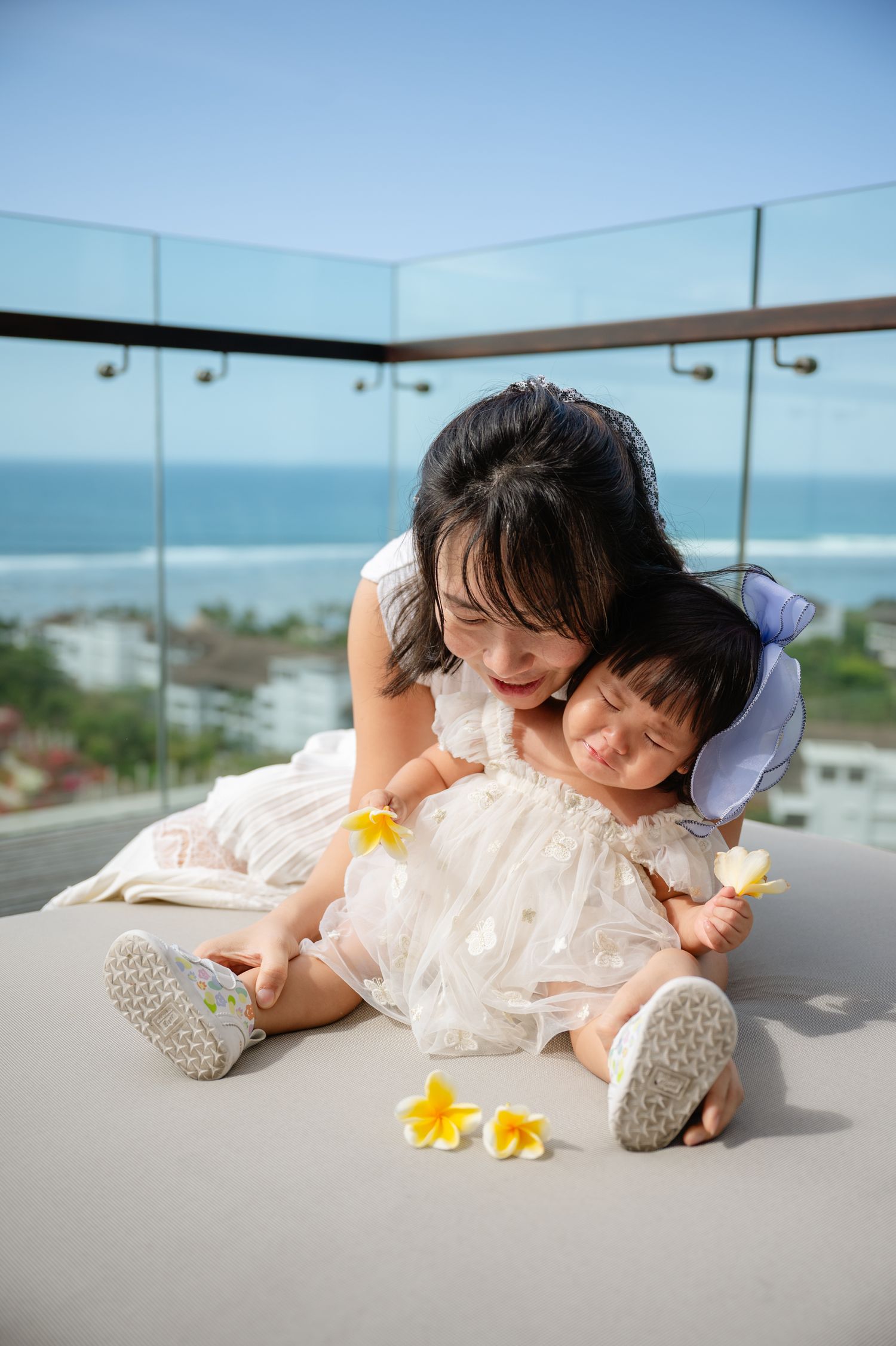 Asian mother hugging baby girl in white dresses on balcony with ocean view, yellow plumeria flowers nearby.