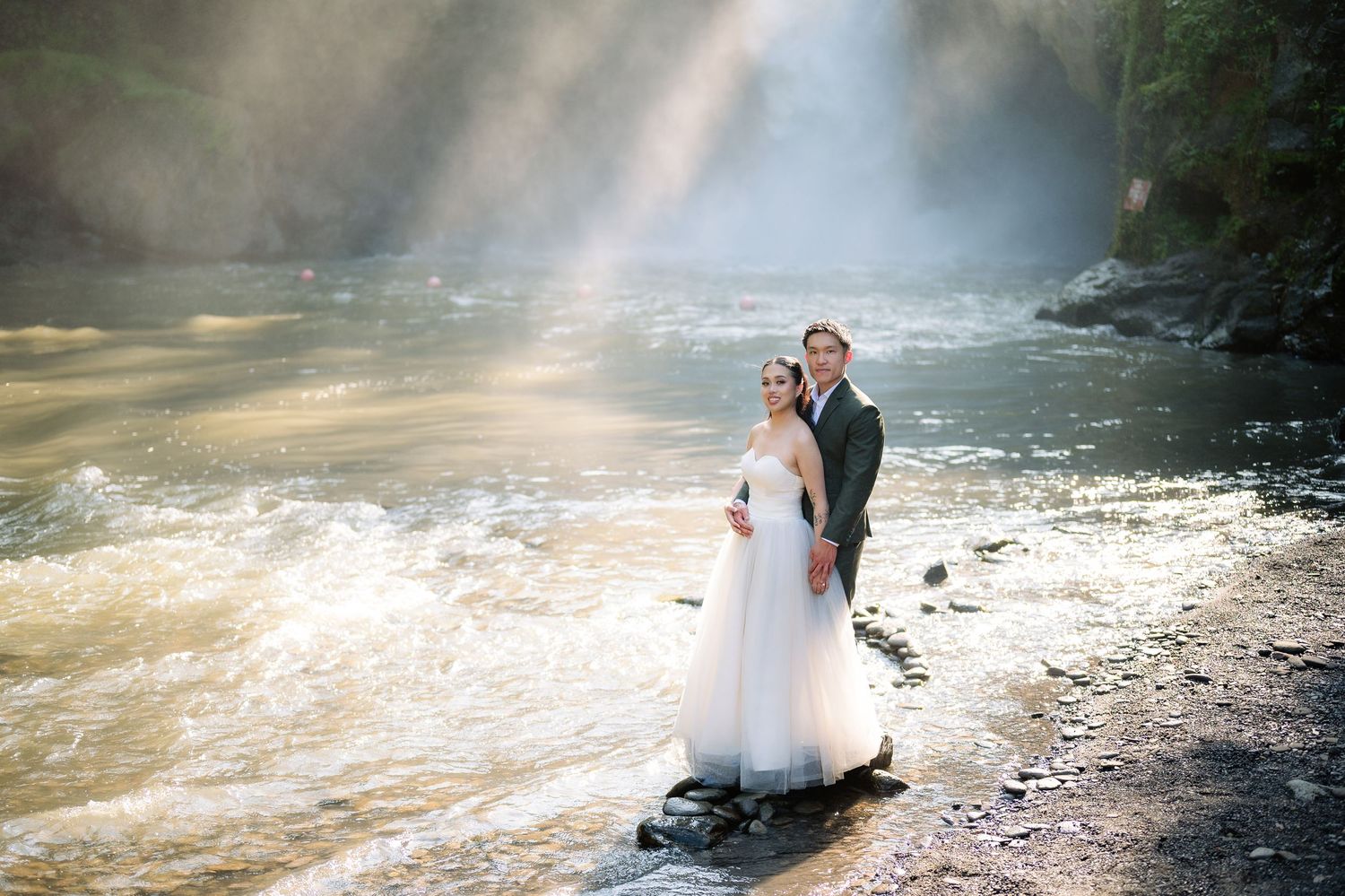 Bride and groom stand on rocks by a sunlit river with a waterfall backdrop on their wedding day.