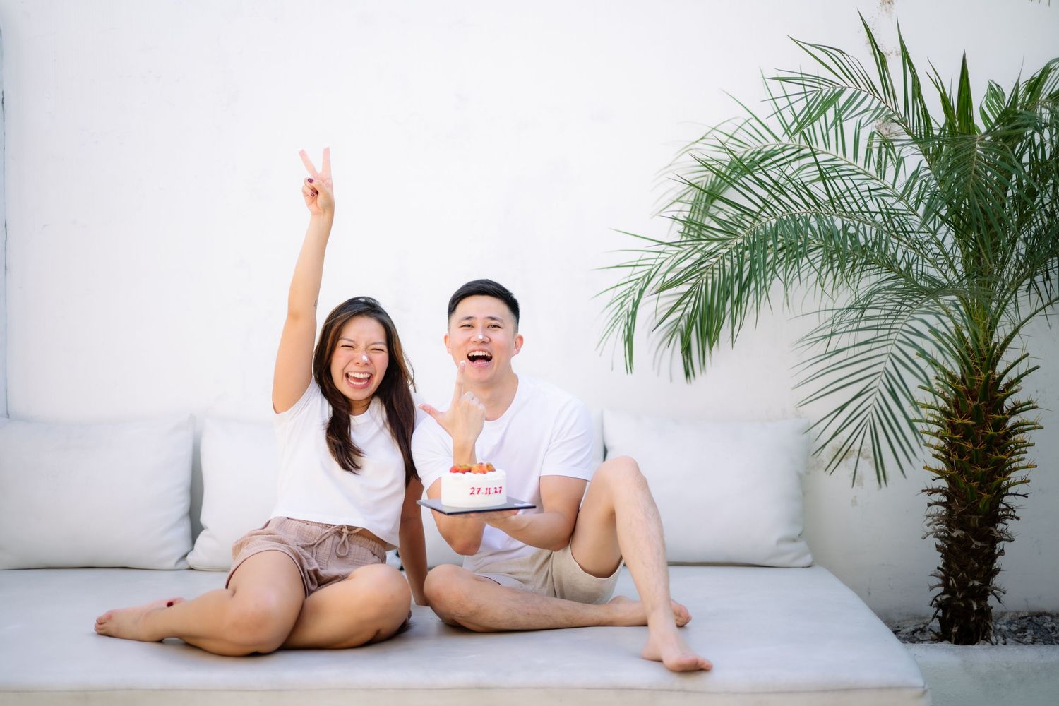 A joyful couple sitting on a white sofa celebrating a birthday with cake, woman raising hand, near a tropical palm plant.