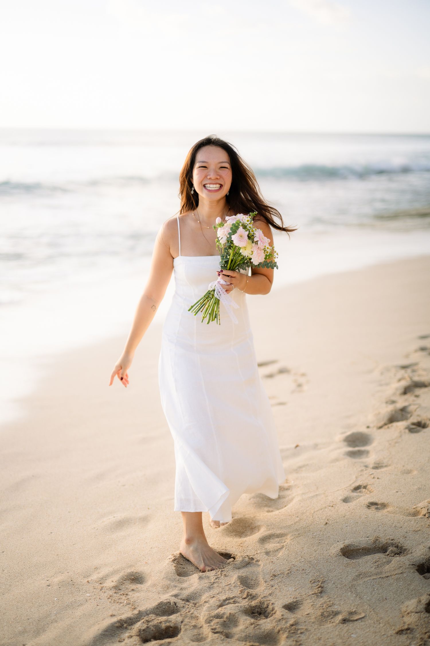 Smiling woman in white dress holding flower bouquet walking barefoot on sandy beach with ocean waves in background.