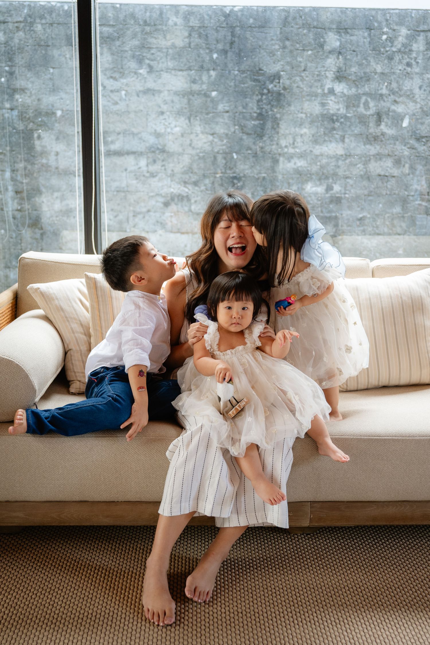 A joyful mother sits on a beige sofa with three young children hugging and laughing together.