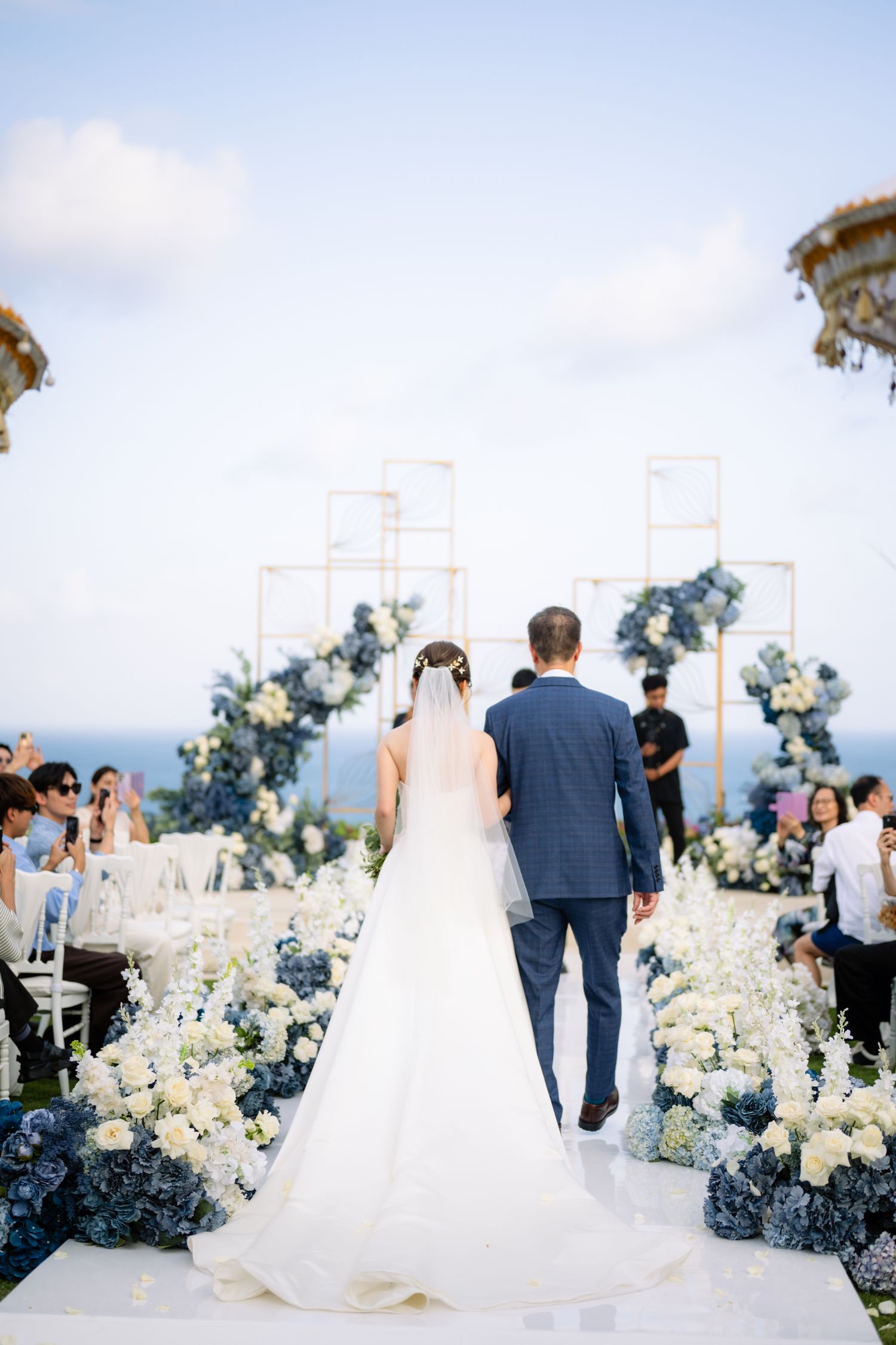 Bride in a long white gown walks the aisle with a man in a blue suit through blue and white flowers at an oceanside wedding.