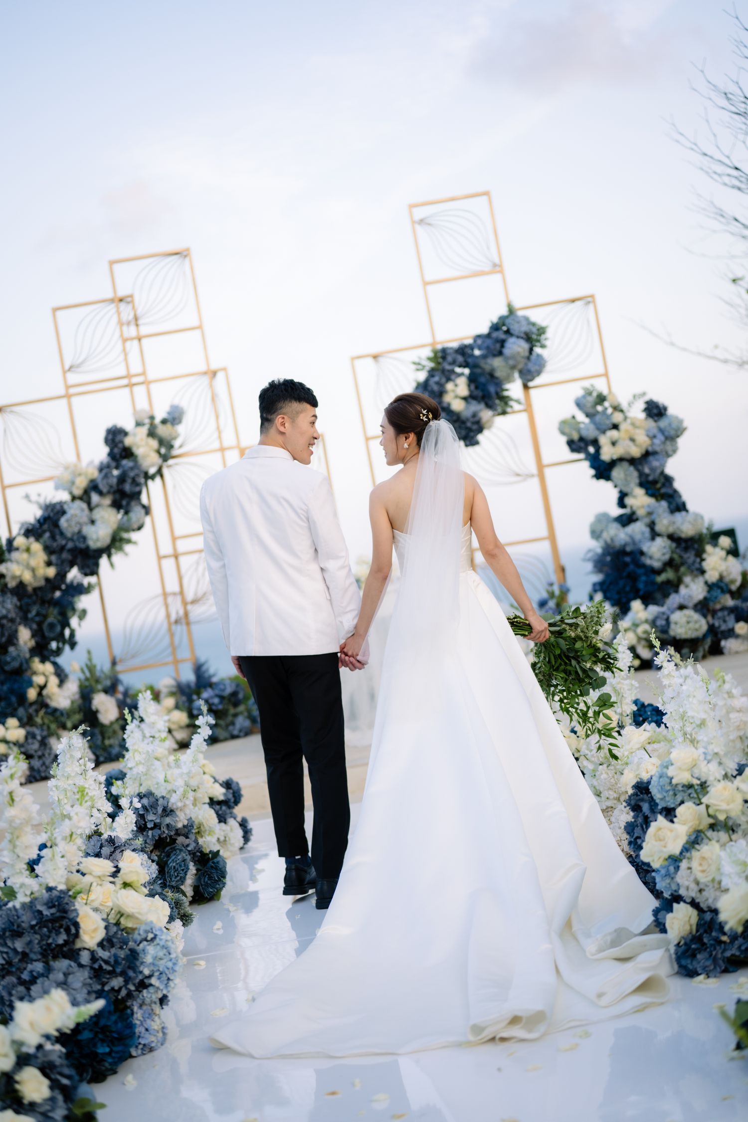 Bride in a flowing white gown and groom in a white jacket hold hands facing a wedding ceremony arch with blue and white floral arrangements.