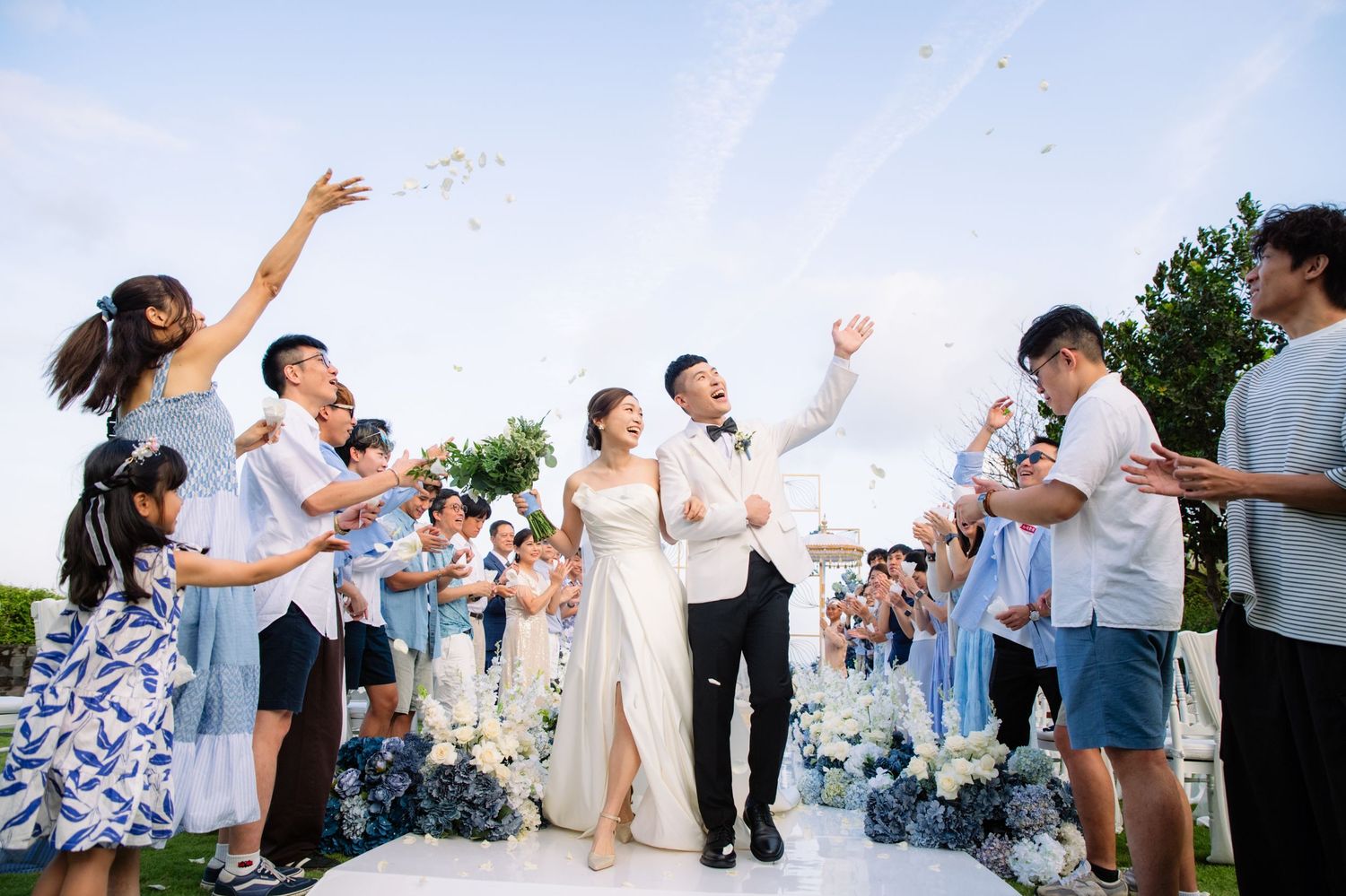 Bride and groom walk down an outdoor aisle as joyful wedding guests toss petals and cheer under a bright blue sky.