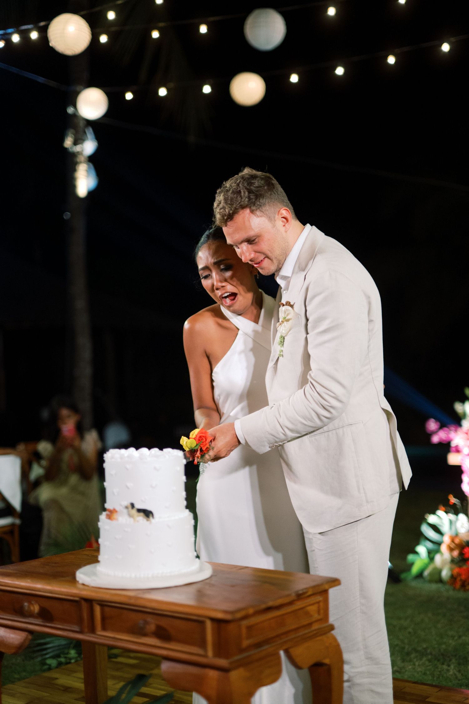 Bride and groom cutting white two-tier wedding cake under string lights at night.