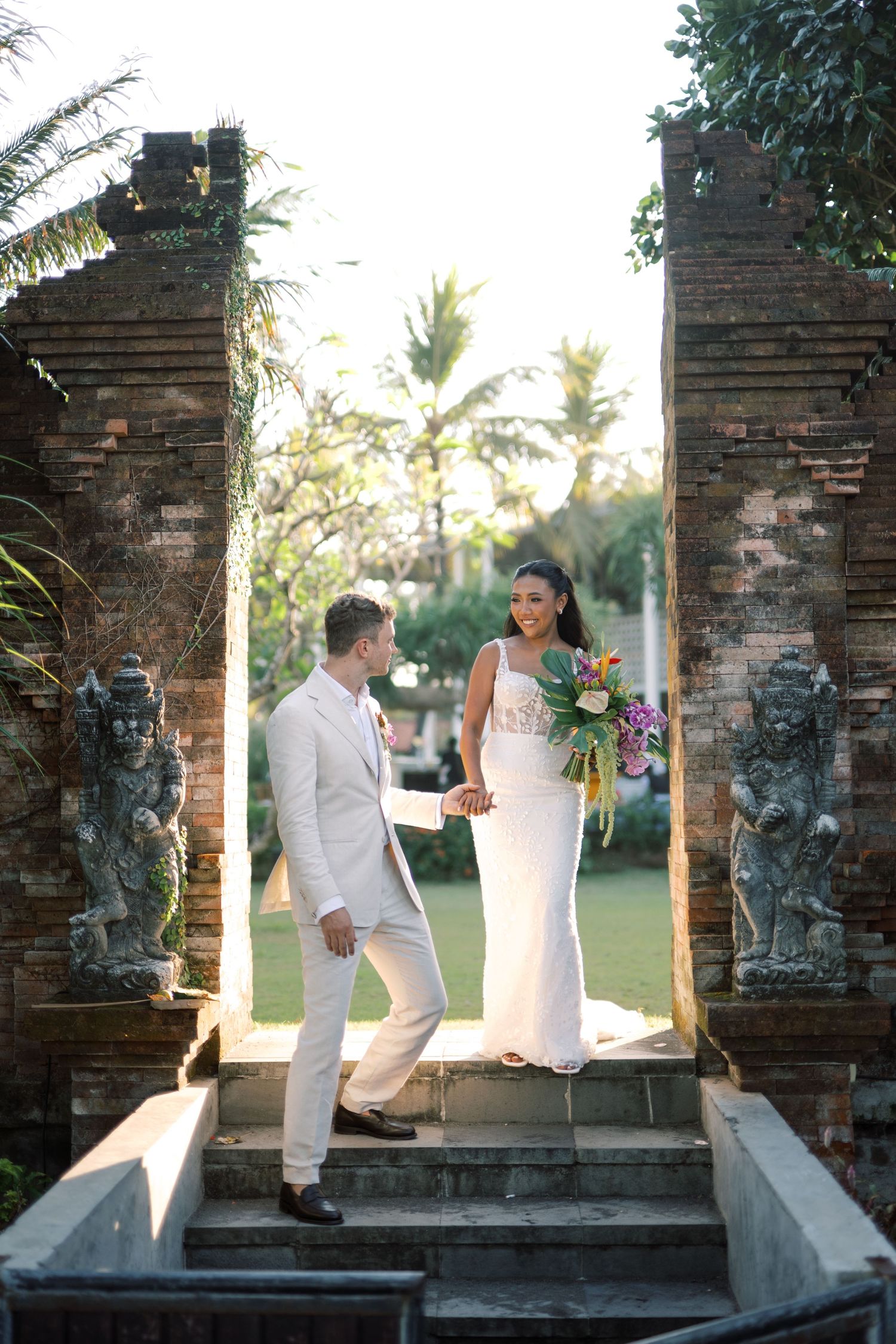 Bride and groom hold hands at ornate Balinese stone gate surrounded by lush tropical greenery.