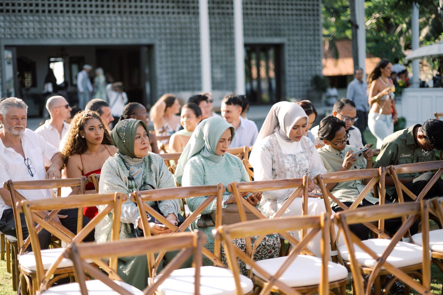 Guests seated outdoors in bamboo chairs at a sunny tropical Bali wedding ceremony.
