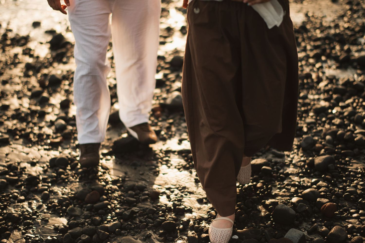 Two people walking hand in hand on a rocky beach, wearing white and brown trousers in warm golden light.