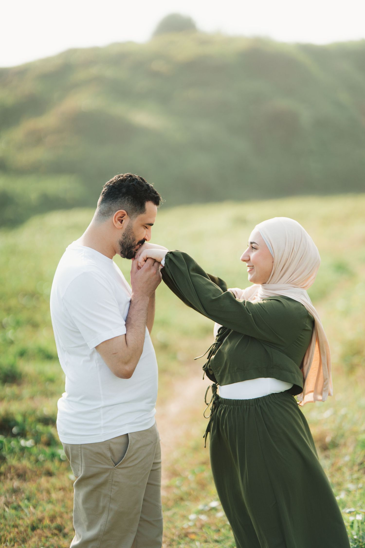 A man kisses a woman's hand outdoors, she wears a green dress and hijab, smiling in a sunlit green field.