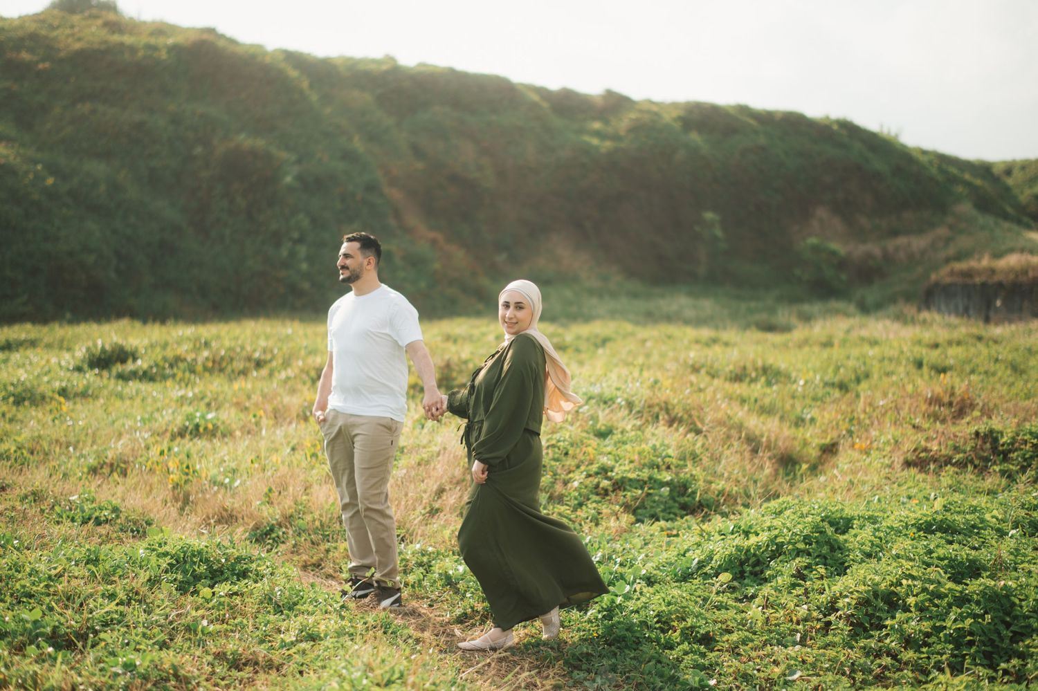 Couple holding hands in a green meadow, woman in hijab and green dress, man in white shirt, golden hour.