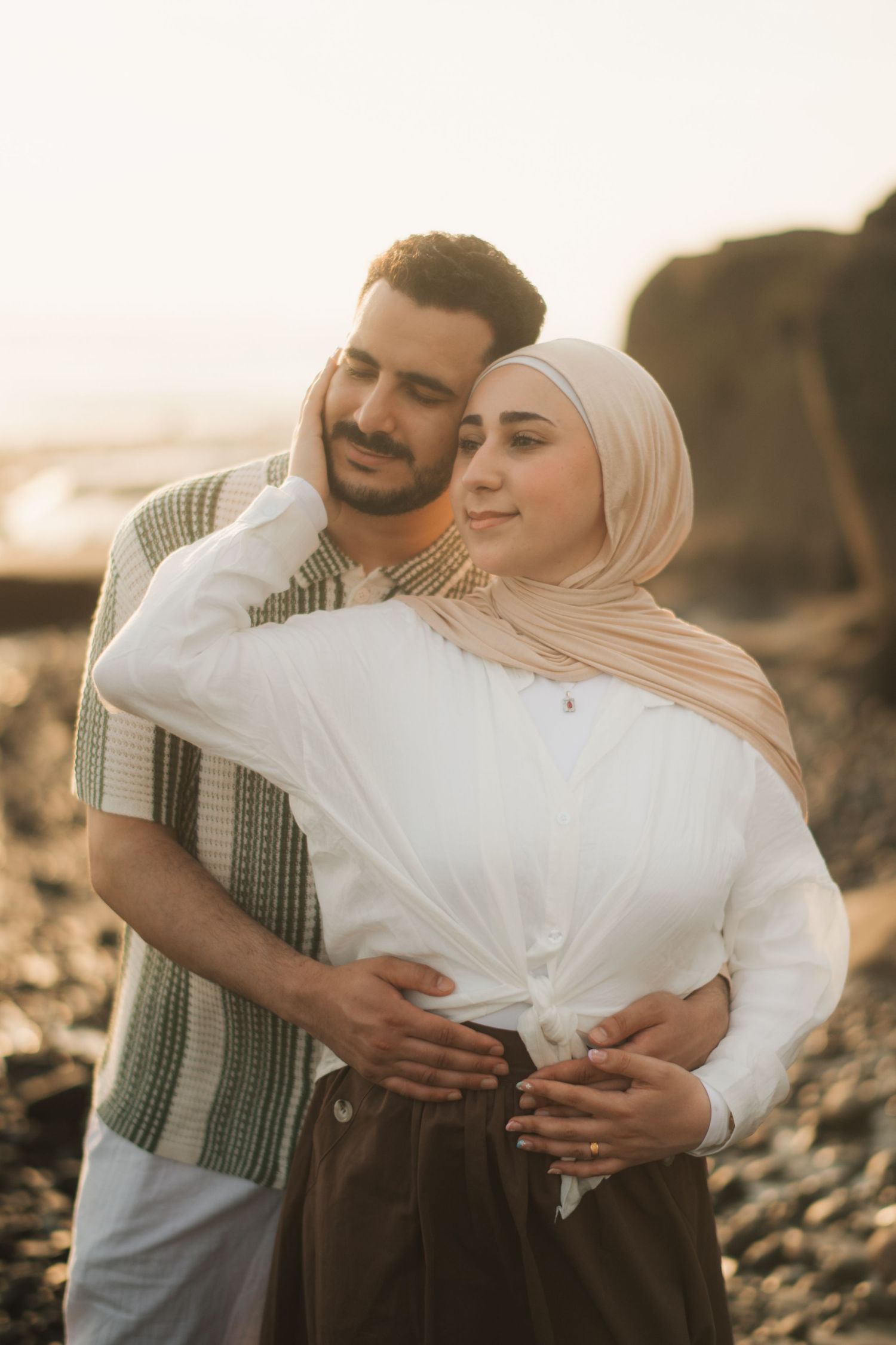 Couple embracing on rocky beach at golden hour, woman in hijab and white top, man in striped shirt.