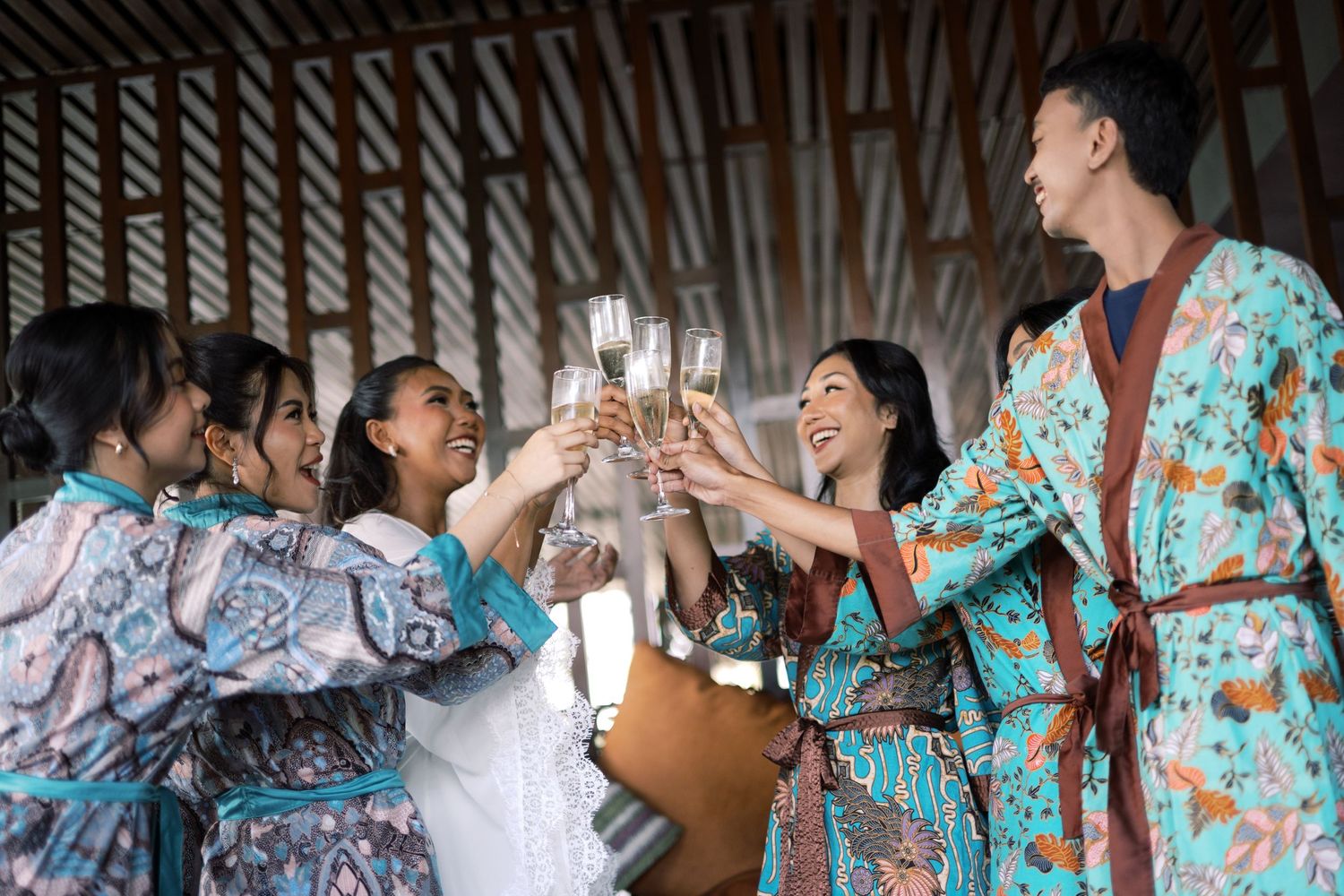 Group of friends in floral robes toasting with champagne glasses at a wedding celebration indoors.