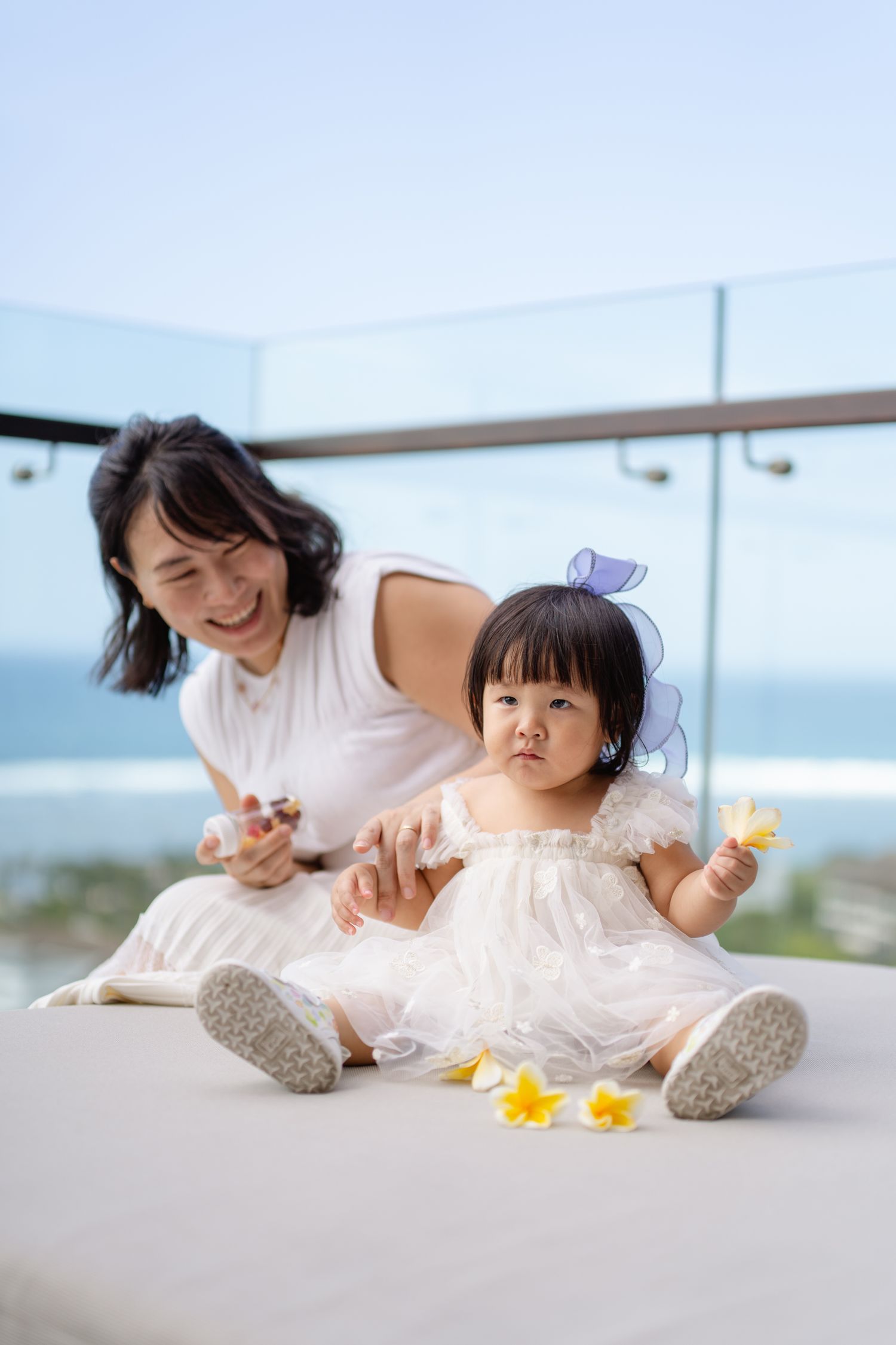 A smiling Asian mother sits with her baby girl in a white dress on a balcony overlooking the ocean.