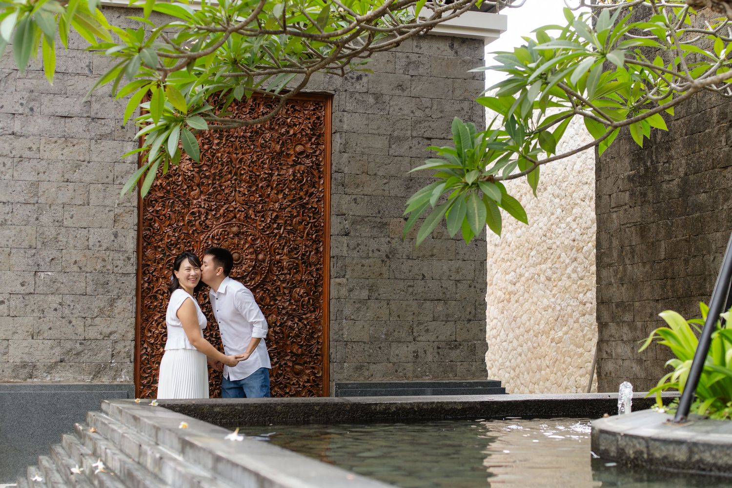 Couple embracing romantically near a tranquil pond surrounded by lush tropical greenery and elegant stone architecture.