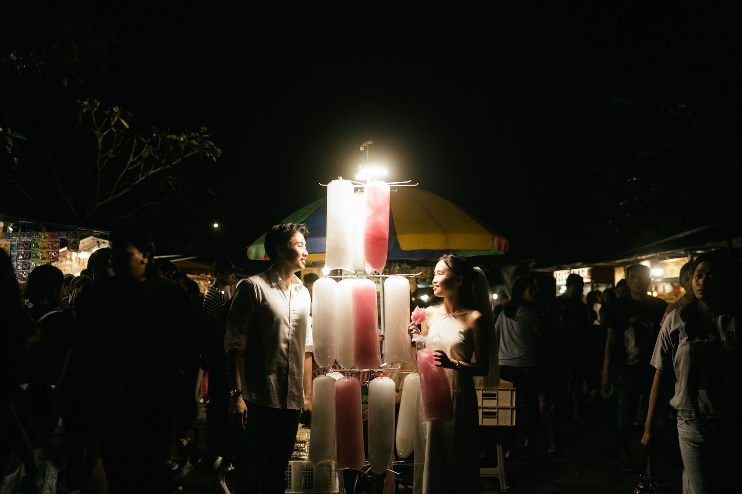 Crowd gathering around illuminated popsicle stand at outdoor night market.
