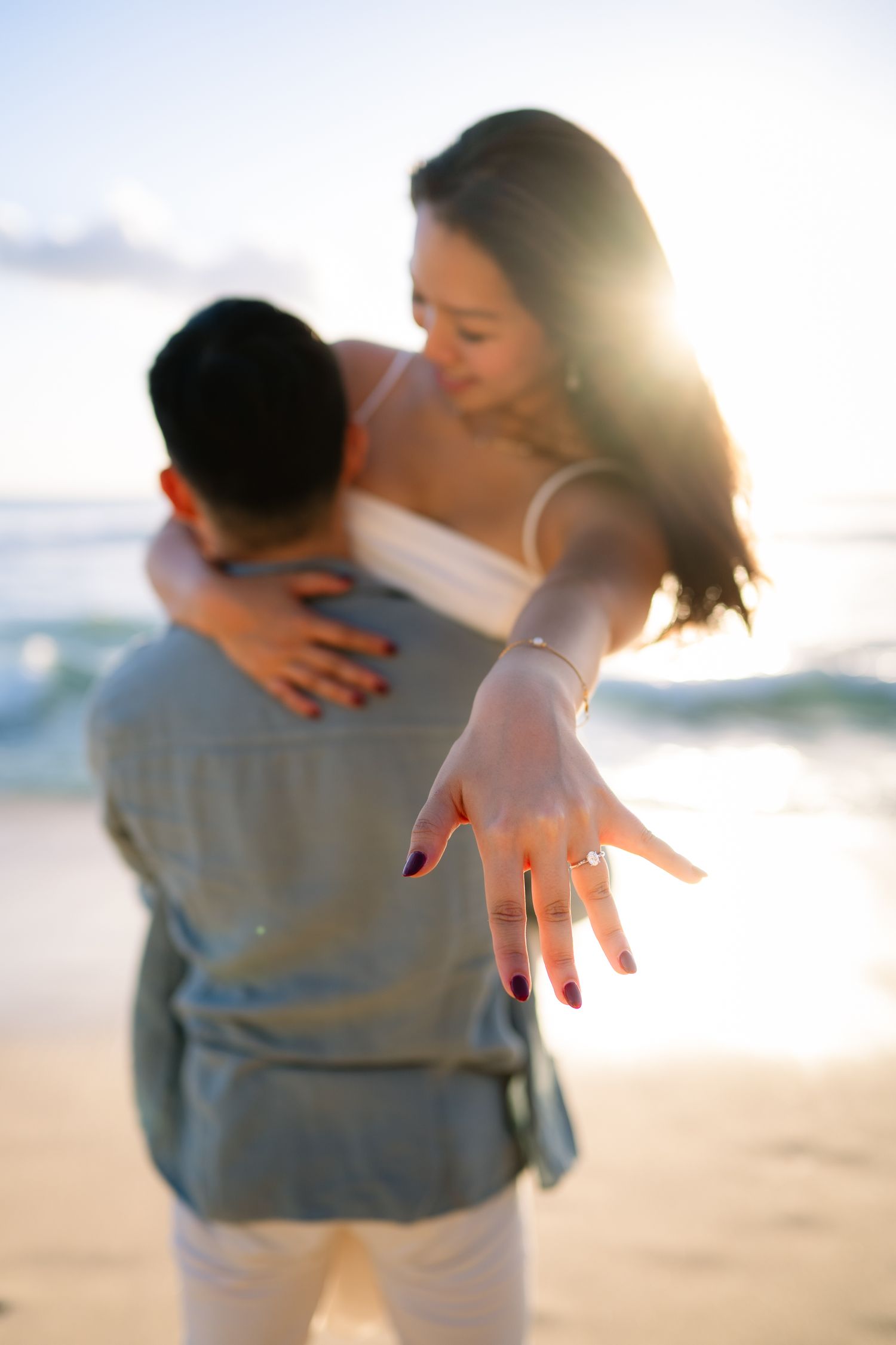 Couple embracing on beach at sunset, woman showing off engagement ring toward camera with ocean backdrop.