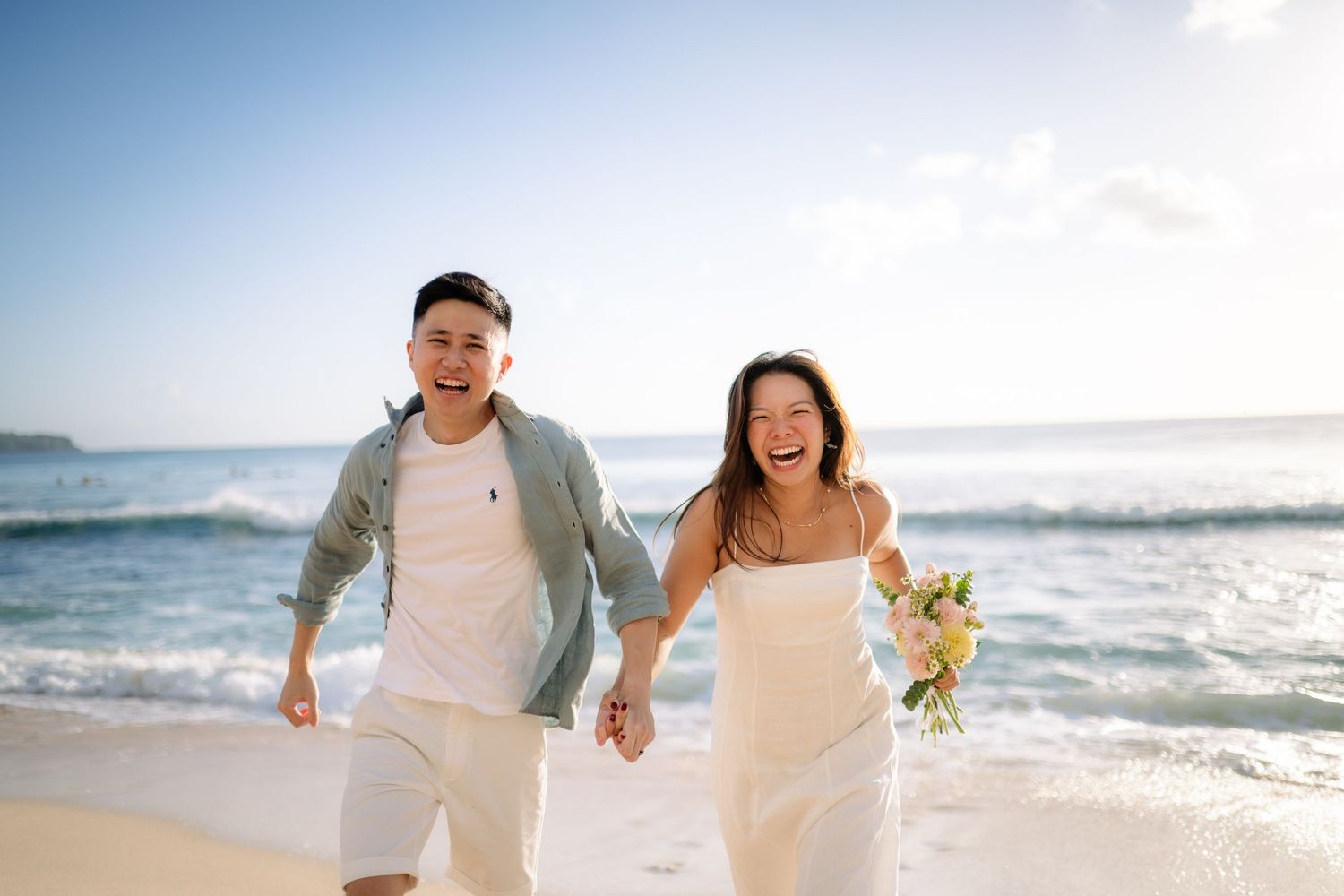 Happy couple in white outfits holding hands and running on a sunny beach, bride carrying a bouquet.
