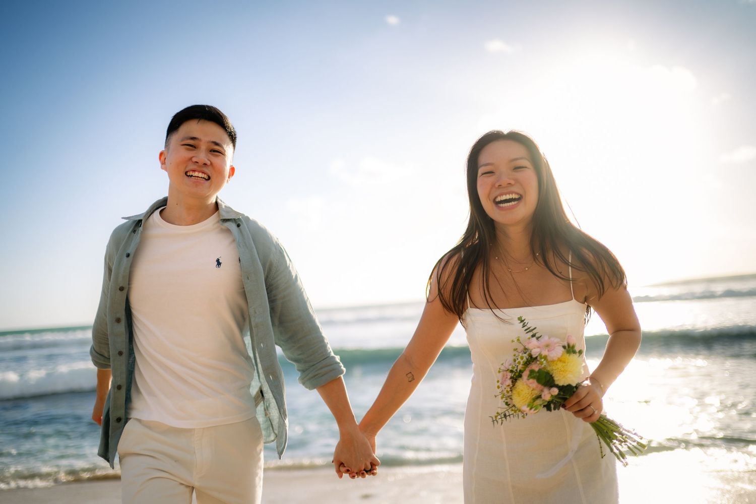 Happy couple holding hands on a sunny beach, bride in white dress holding yellow flowers, groom in casual attire.