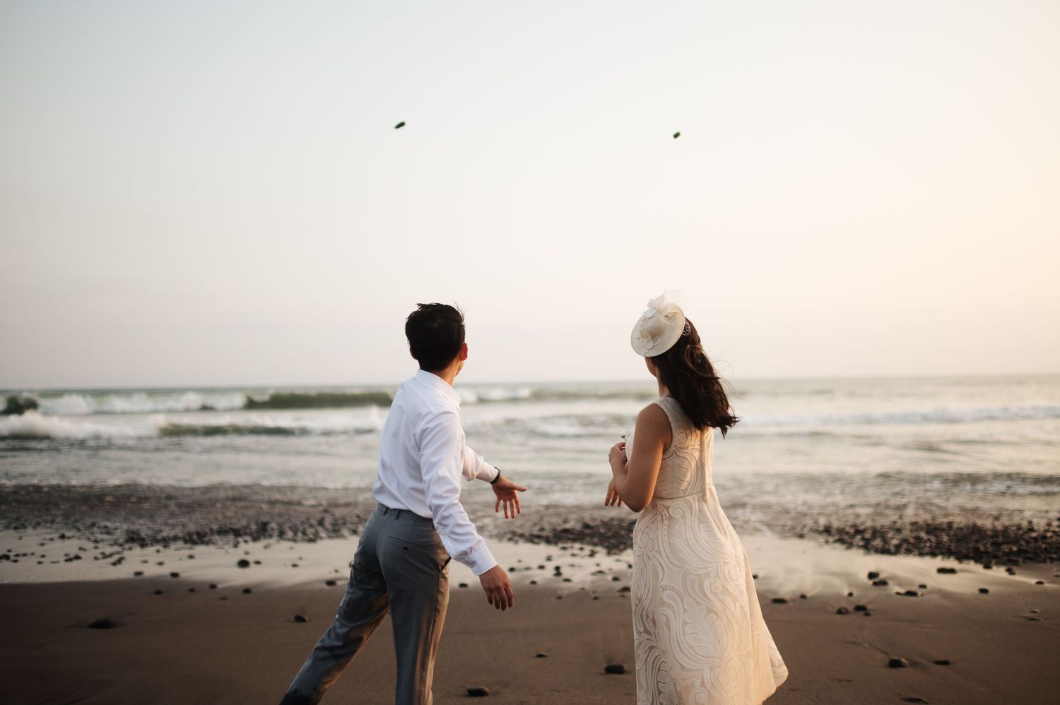 Couple in wedding attire holding hands on a sandy beach at sunset with ocean waves in the background.
