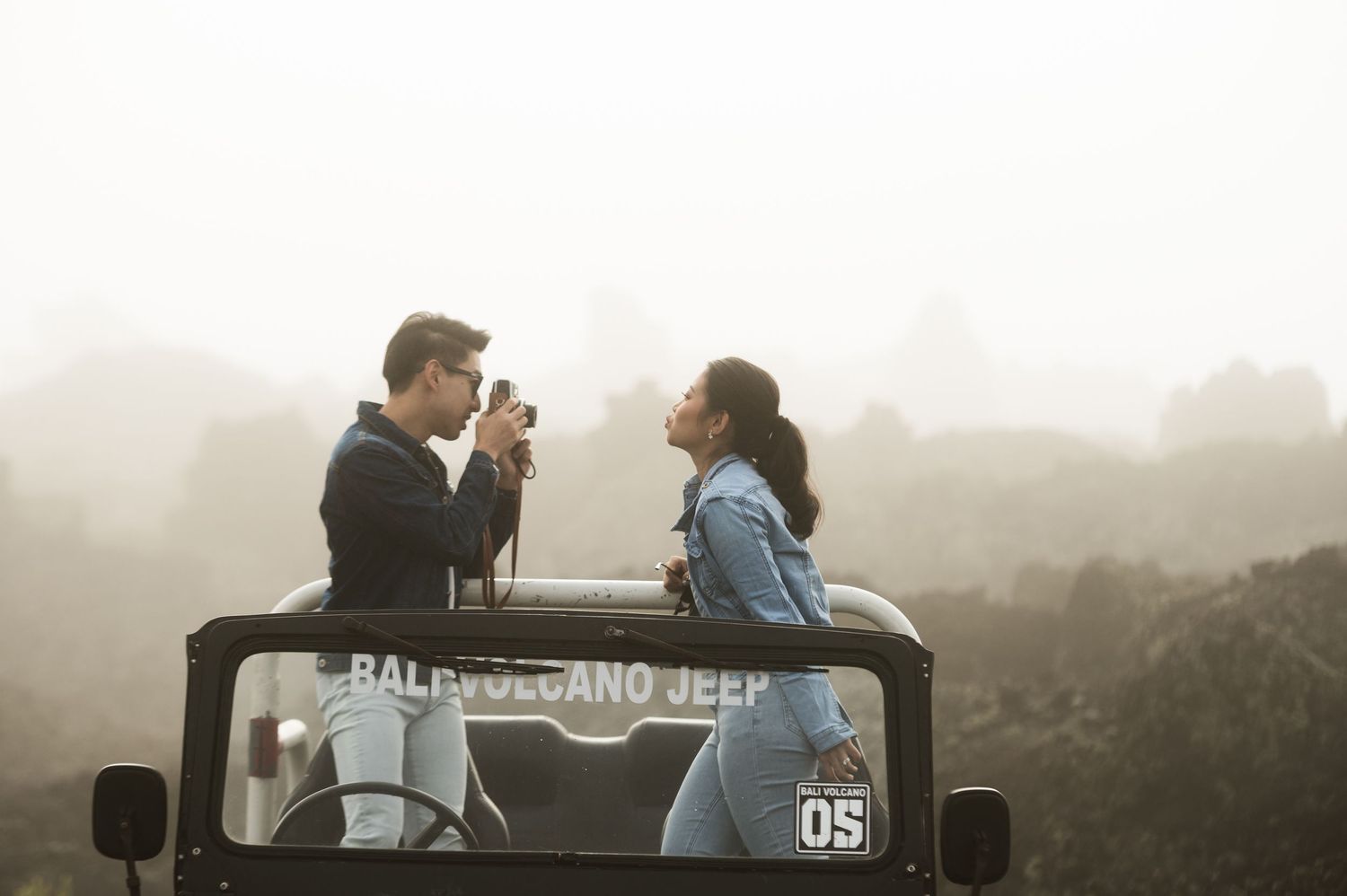 Couple sitting on a Bali Volcano Jeep in misty mountains, man photographing woman in romantic outdoor adventure.
