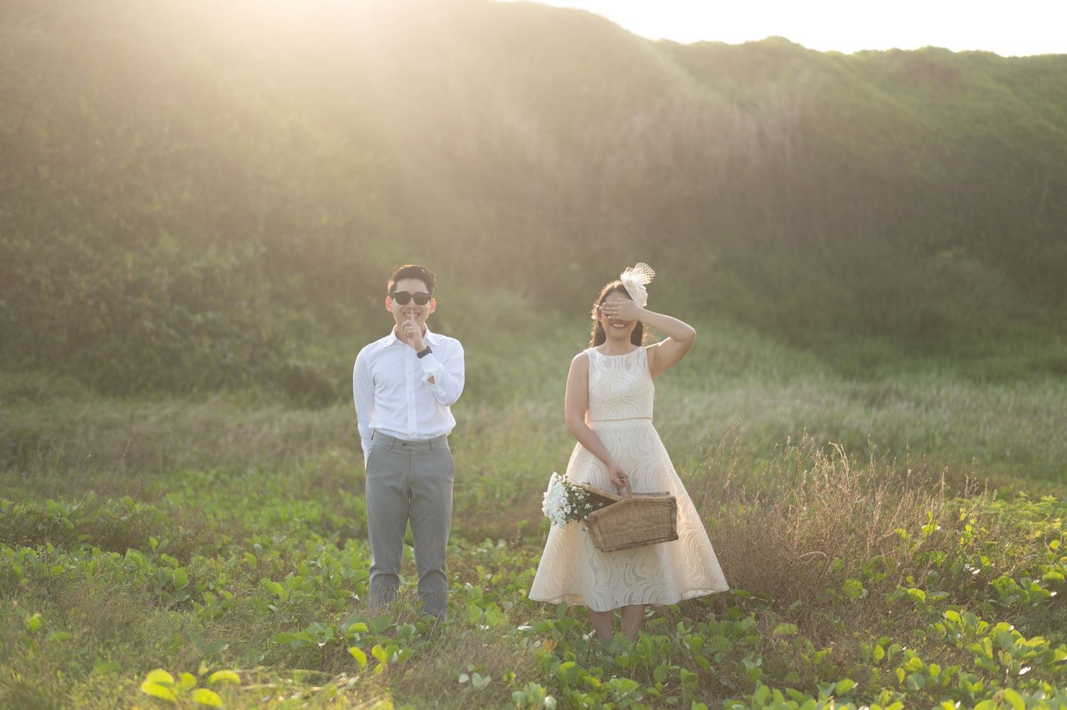 A young boy and girl in formal attire stand in a sunlit green field, girl holding a basket.