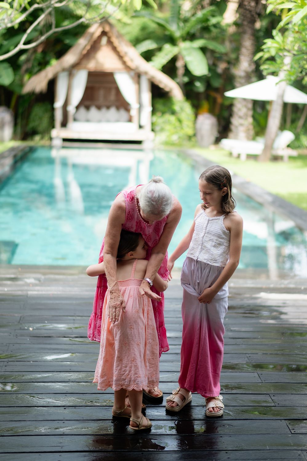 Grandmother bending down to hug young girl while another child stands nearby by a tropical pool.