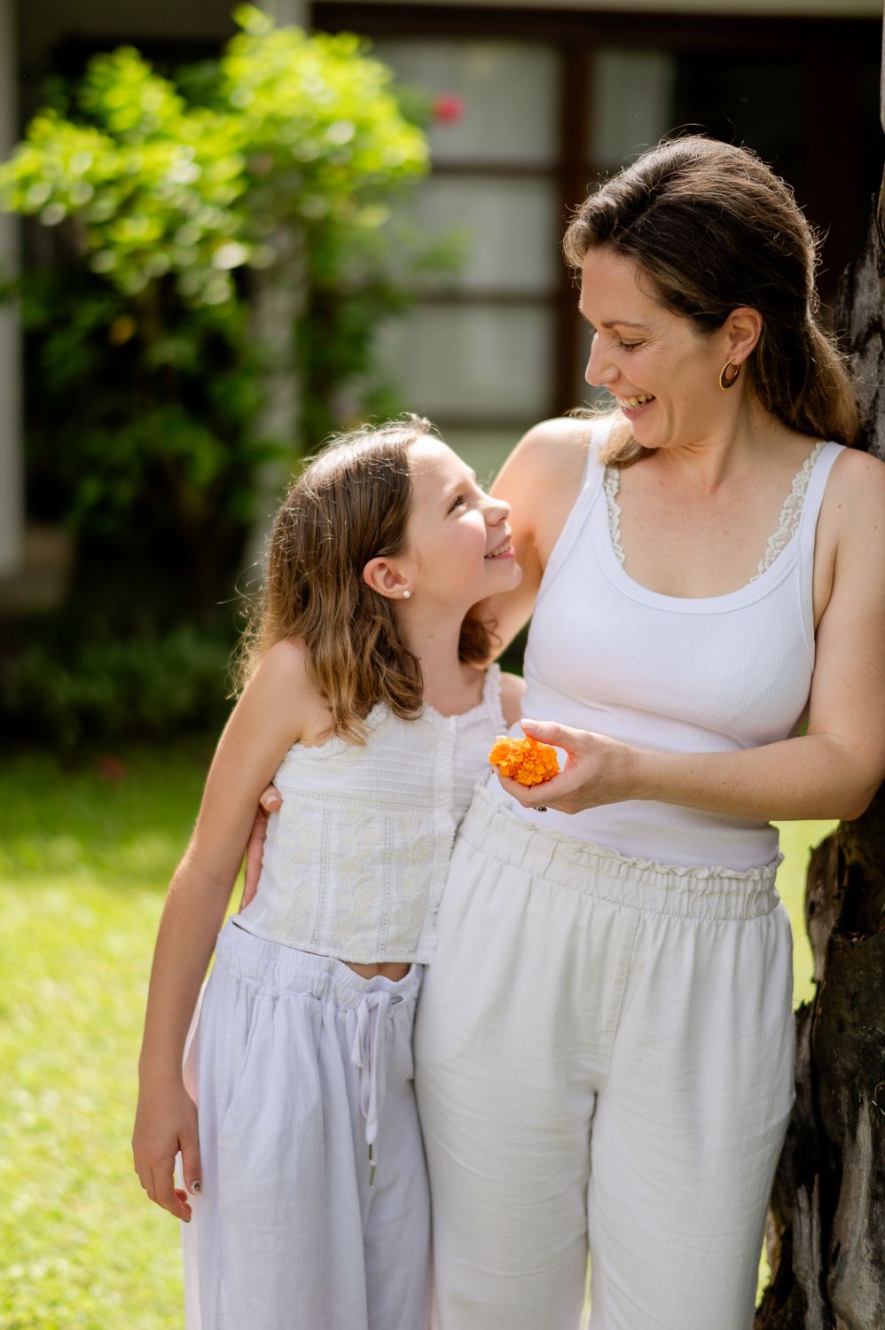 A smiling mother and daughter share a joyful moment outdoors, with the woman holding orange flowers near a tree.