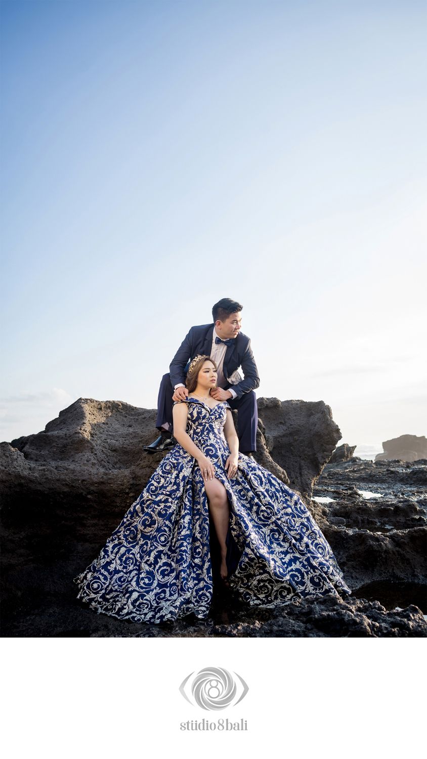 Couple in elegant blue patterned gown and navy suit posing on rocky coastal cliffs for a pre-wedding photoshoot.